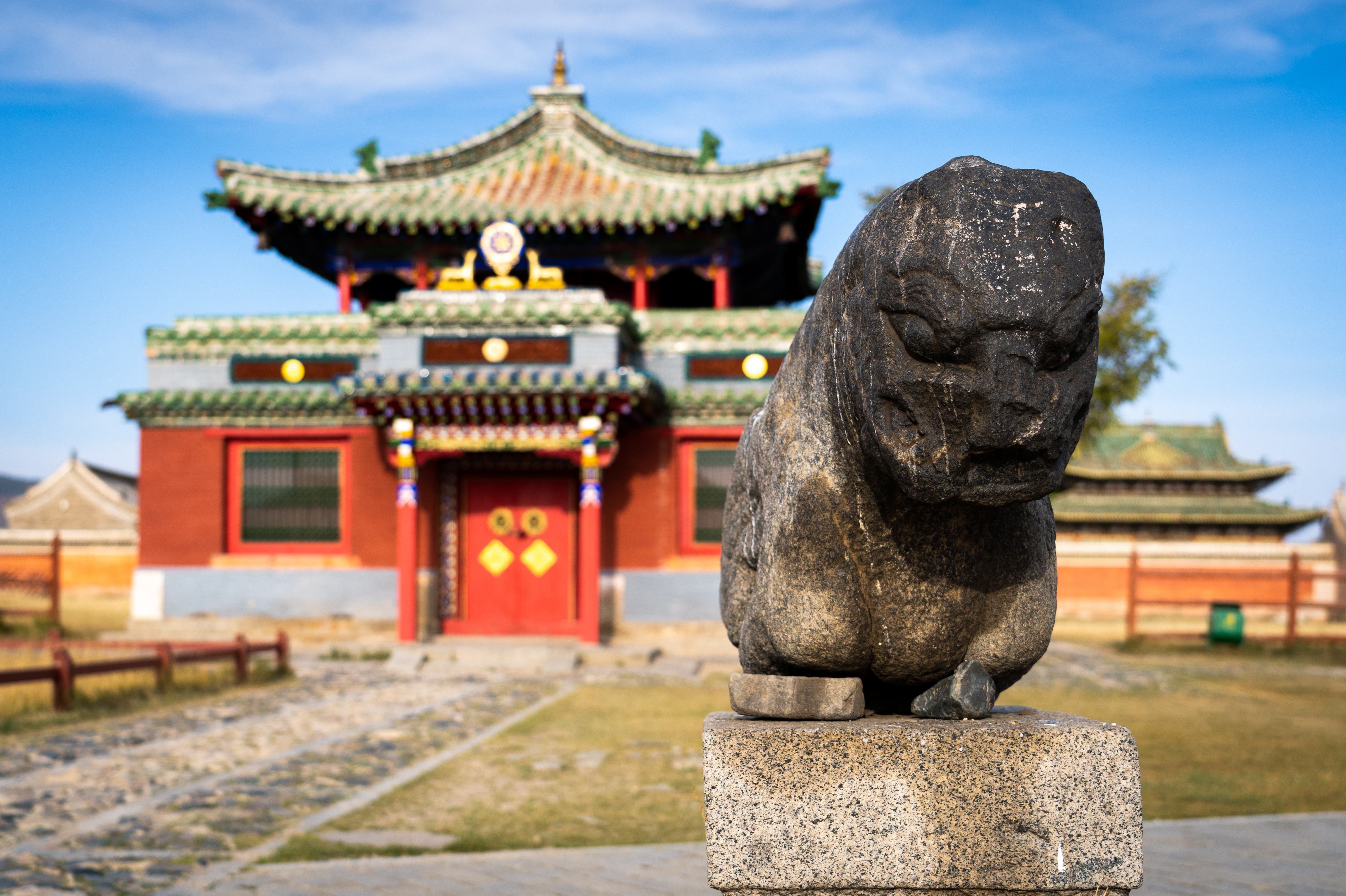 Weathered stone lion statue on a plinth before a red and green temple building at Erdene Zuu Monastery in Kharkhorin, Mongolia