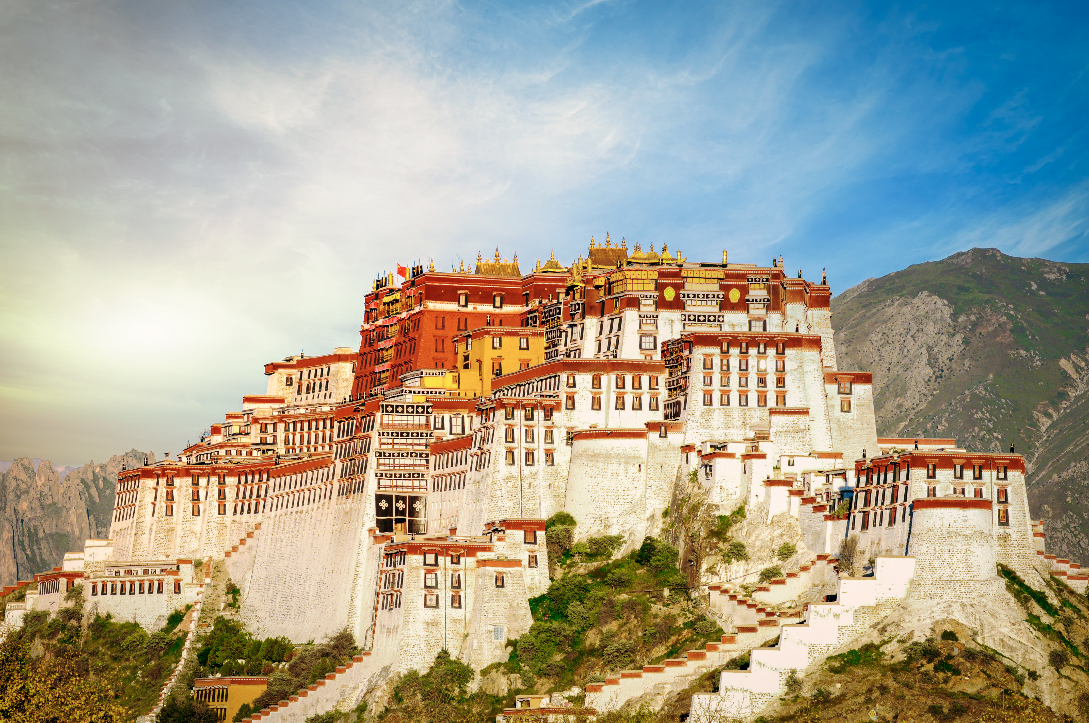 Potala Palace rises in tiered white and red stone walls up a hillside in Lhasa, its golden roofs catching the light against a mountain backdrop.
