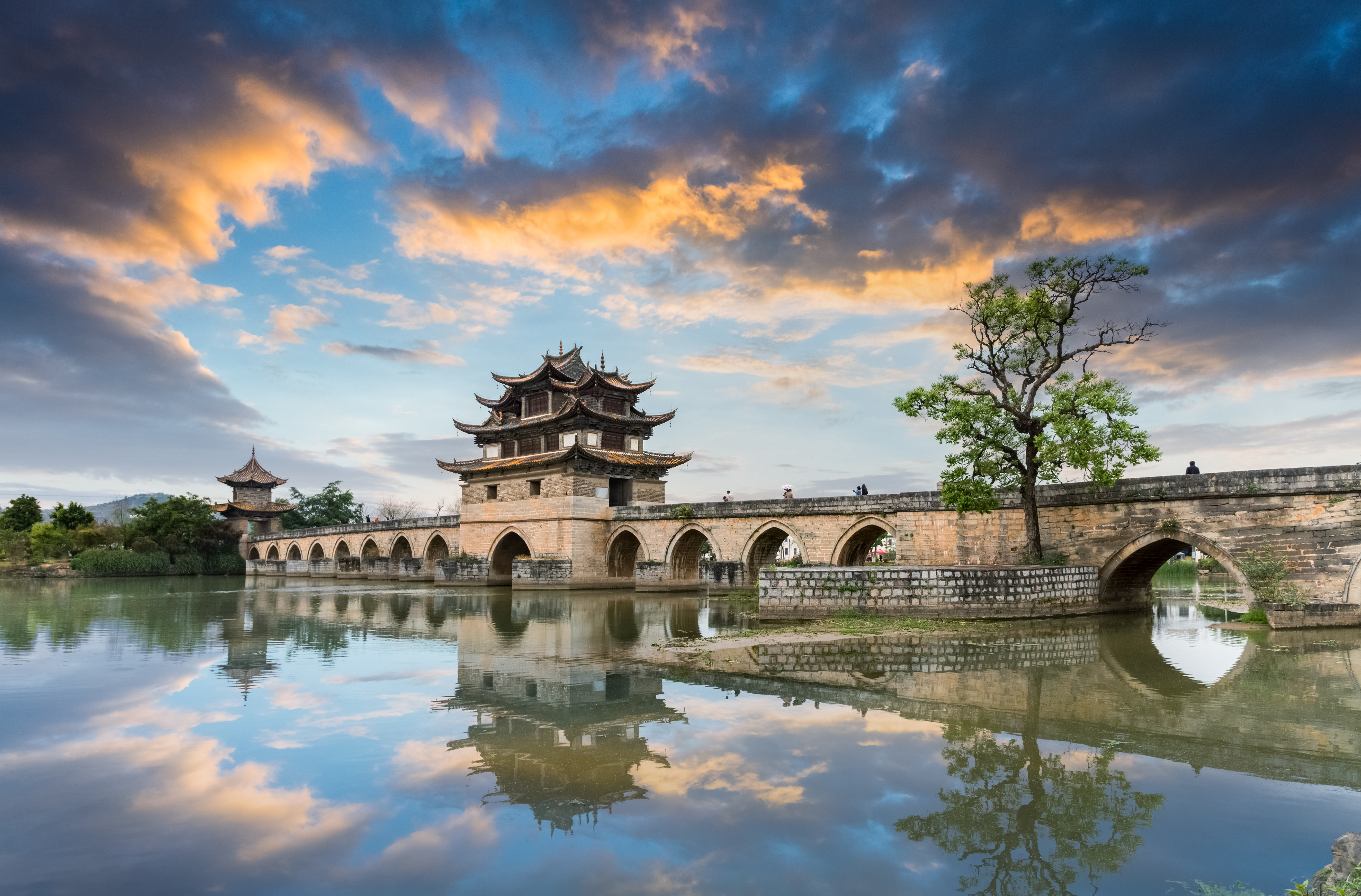 Multi-tiered pavilion bridge with arched stone spans reflected in still water at sunset, near Jianshui, Yunnan