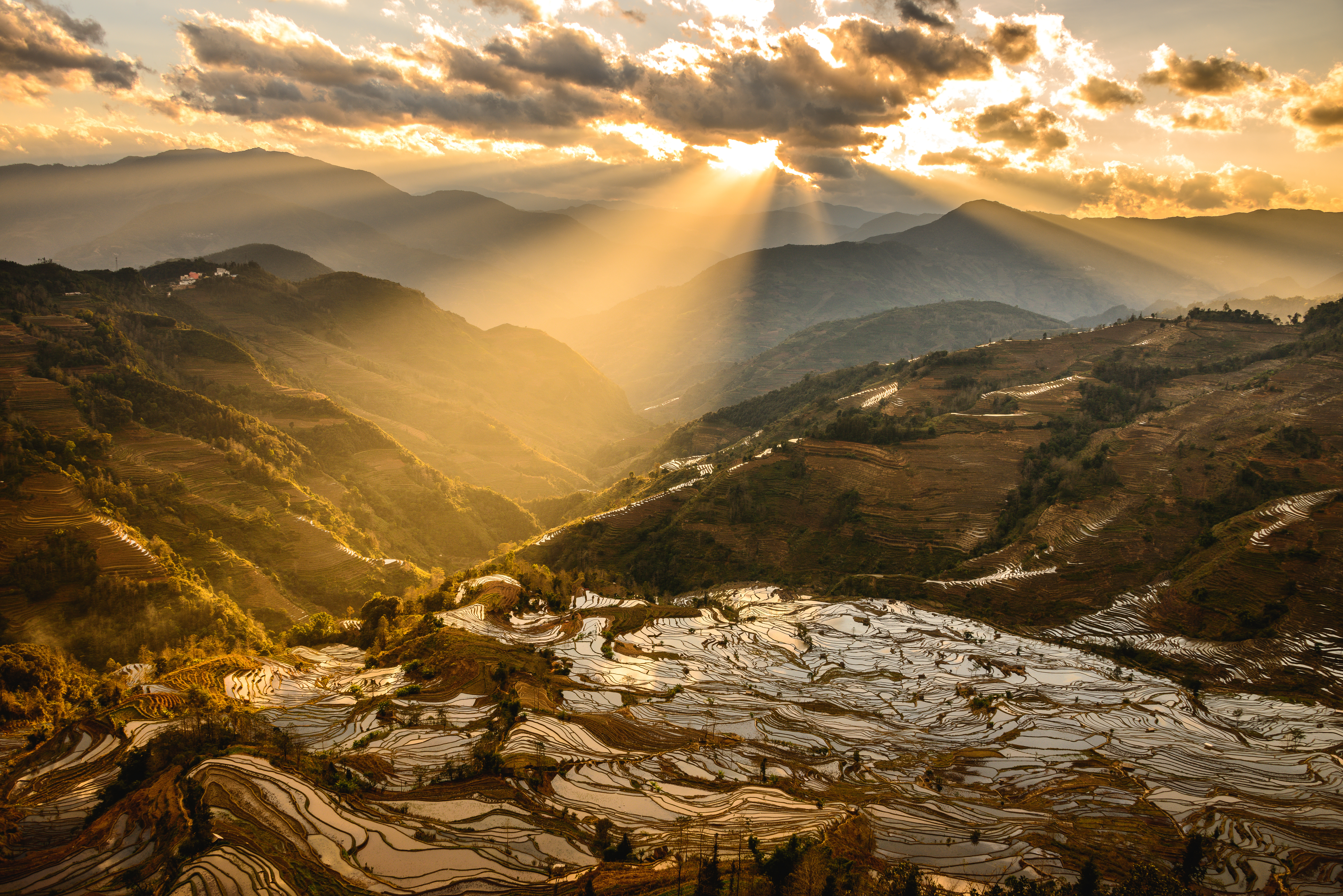 Sun rays break through clouds over flooded rice terraces at Tiger Mouth, Yuanyang, with layered mountains fading into golden haze