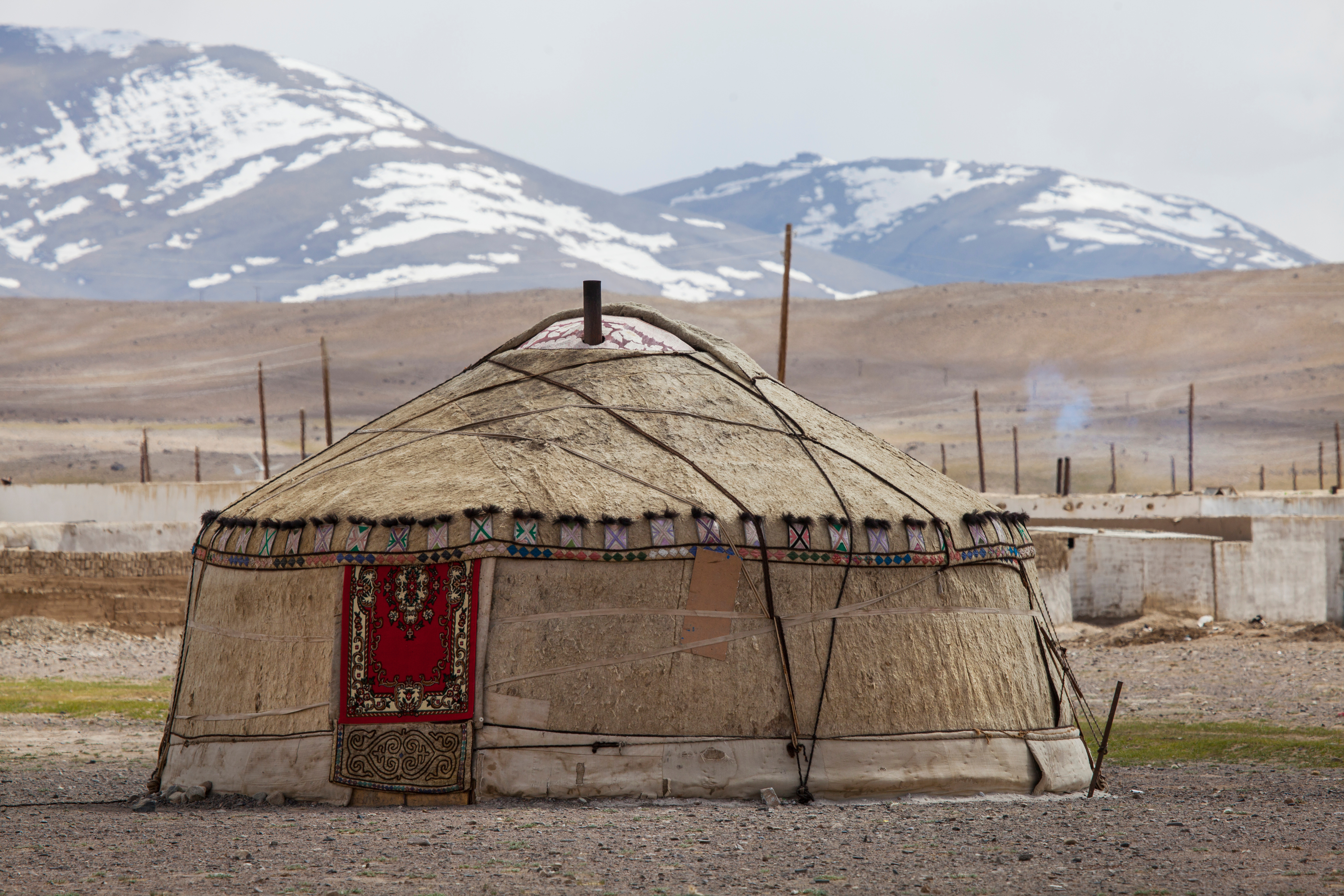 A traditional felt yurt with a red patterned door rug stands before snow-capped peaks near Karakul Lake, Tajikistan