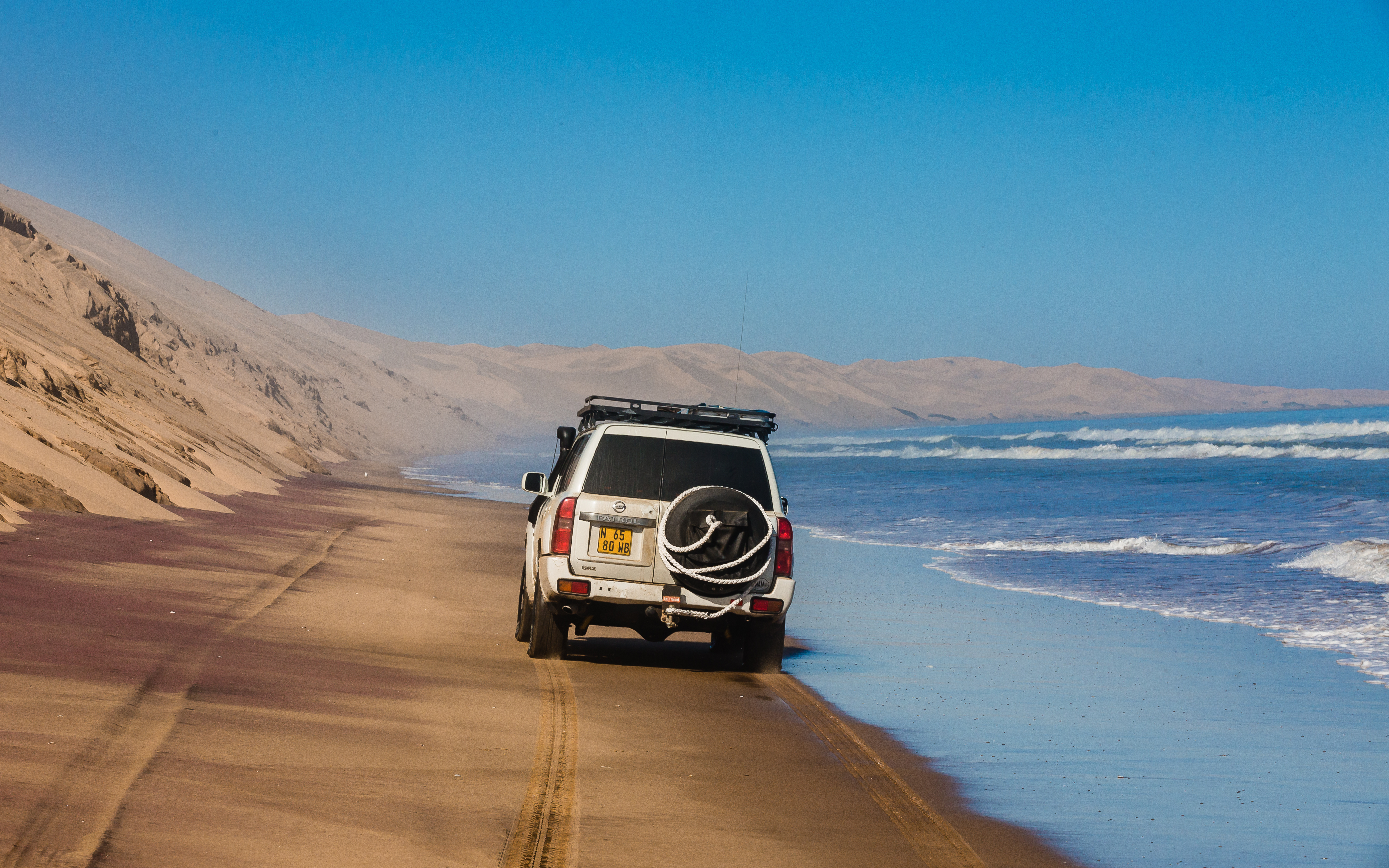 White 4x4 with roof rack and spare tire drives along a sandy beach at Walvis Bay, Namibia, dunes on one side and the Atlantic on the other