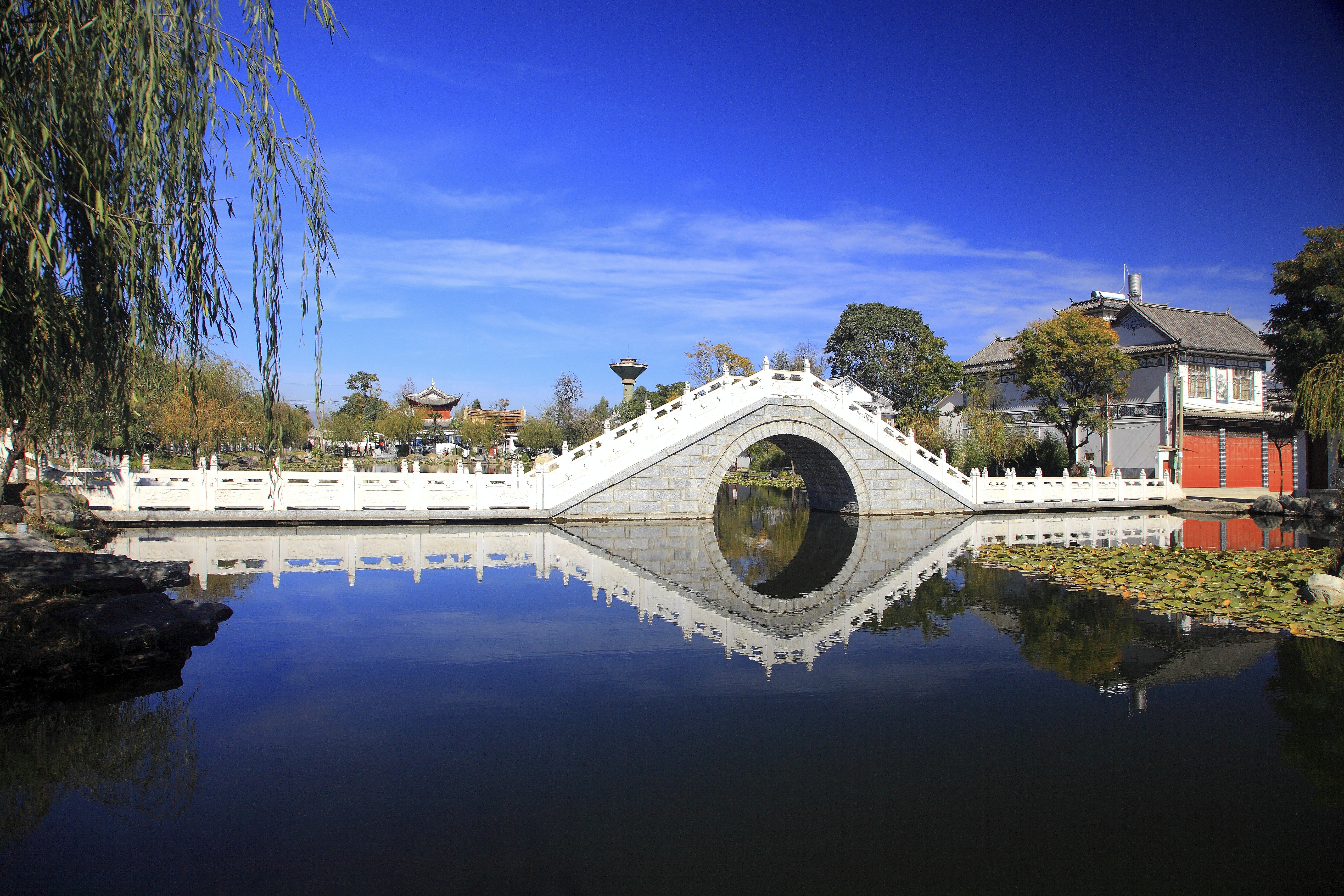 White stone arch bridge reflected in still water, Xizhou town, Yunnan, with willow trees and traditional courtyard buildings nearby