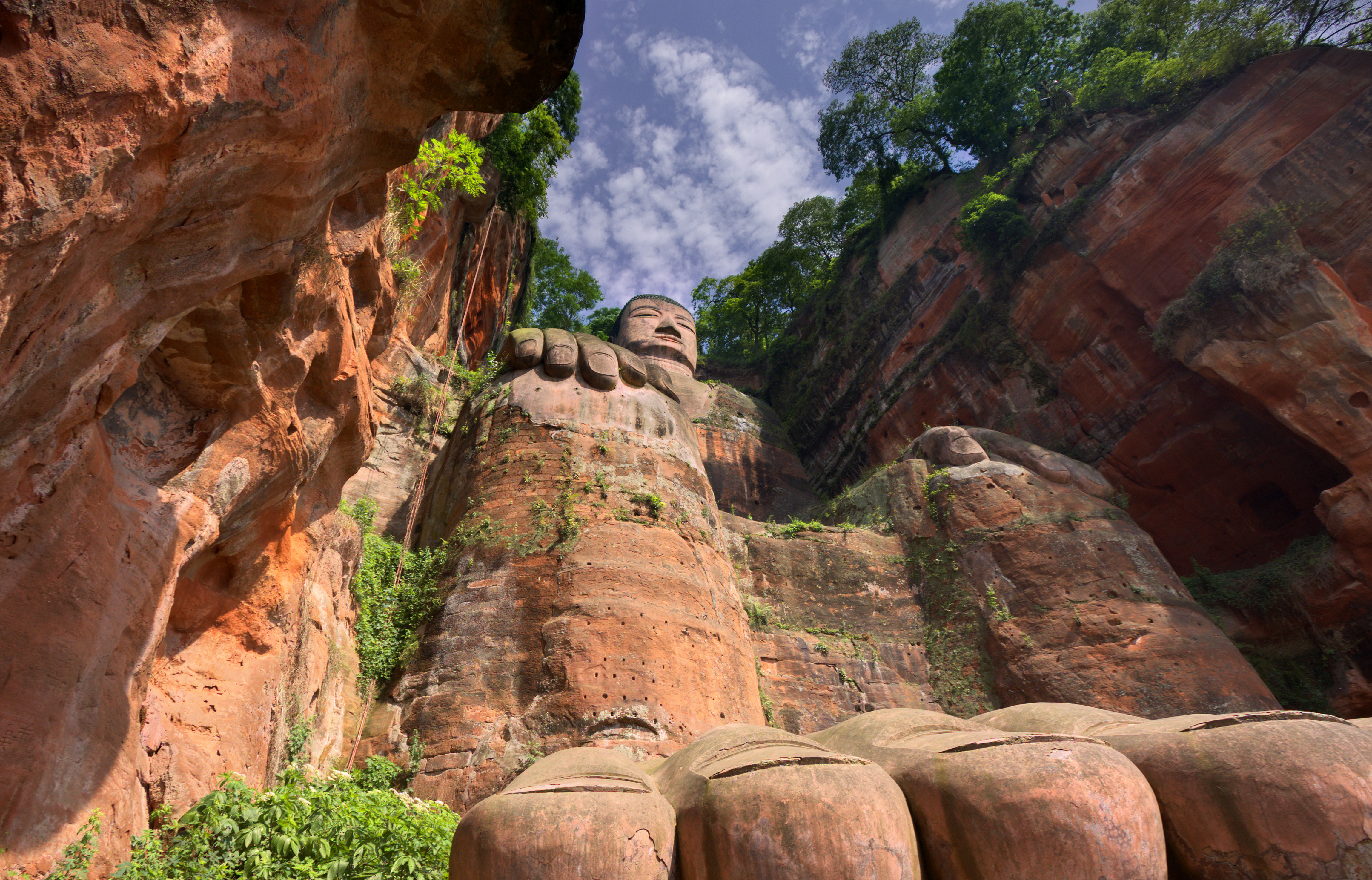 Stone carved head and shoulders of the Leshan Giant Buddha viewed from below, flanked by red sandstone cliffs and green trees against a blue sky