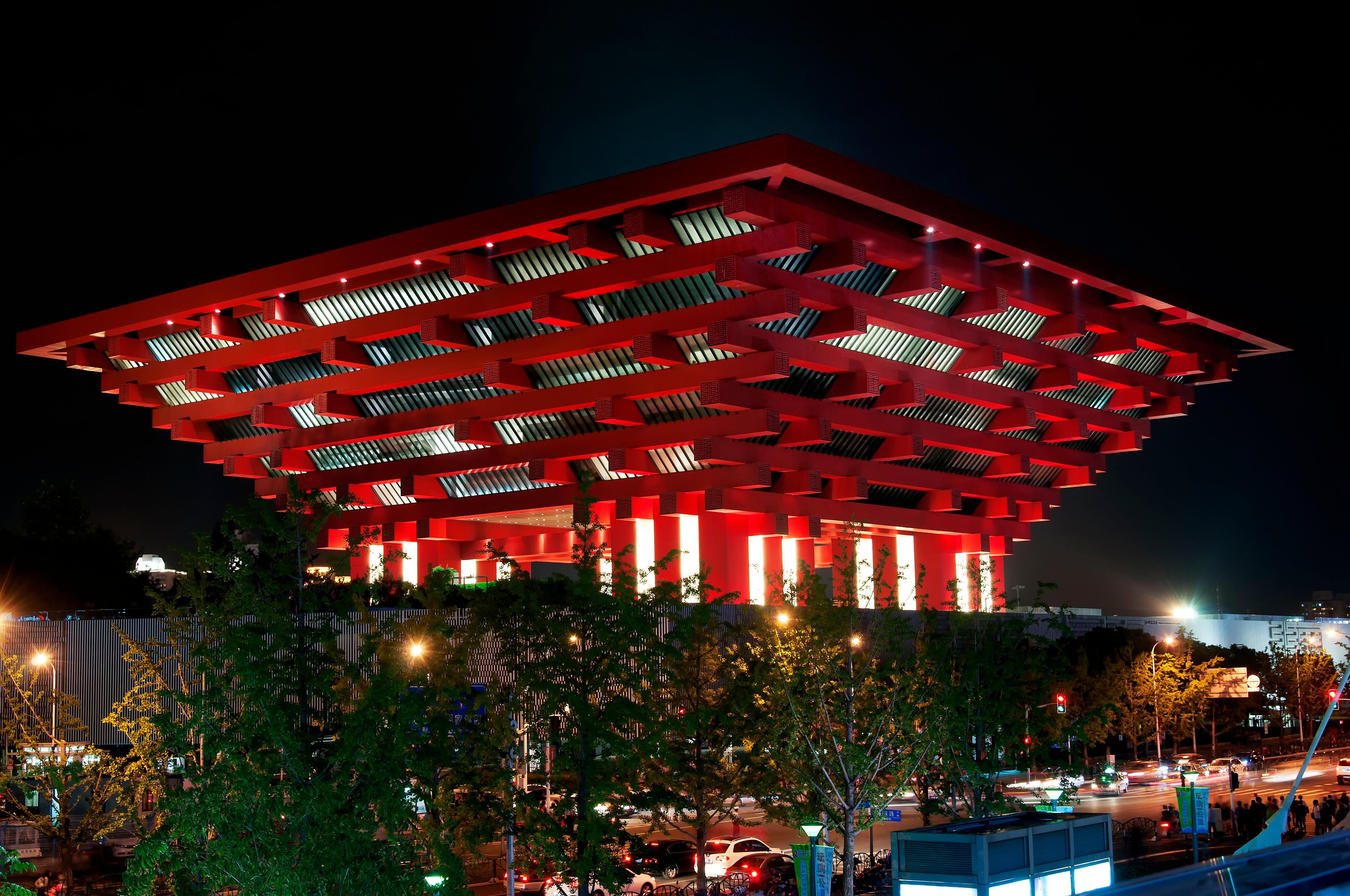 The China Art Museum in Shanghai illuminated in red at night, its tiered inverted pyramid form rising above a row of trees