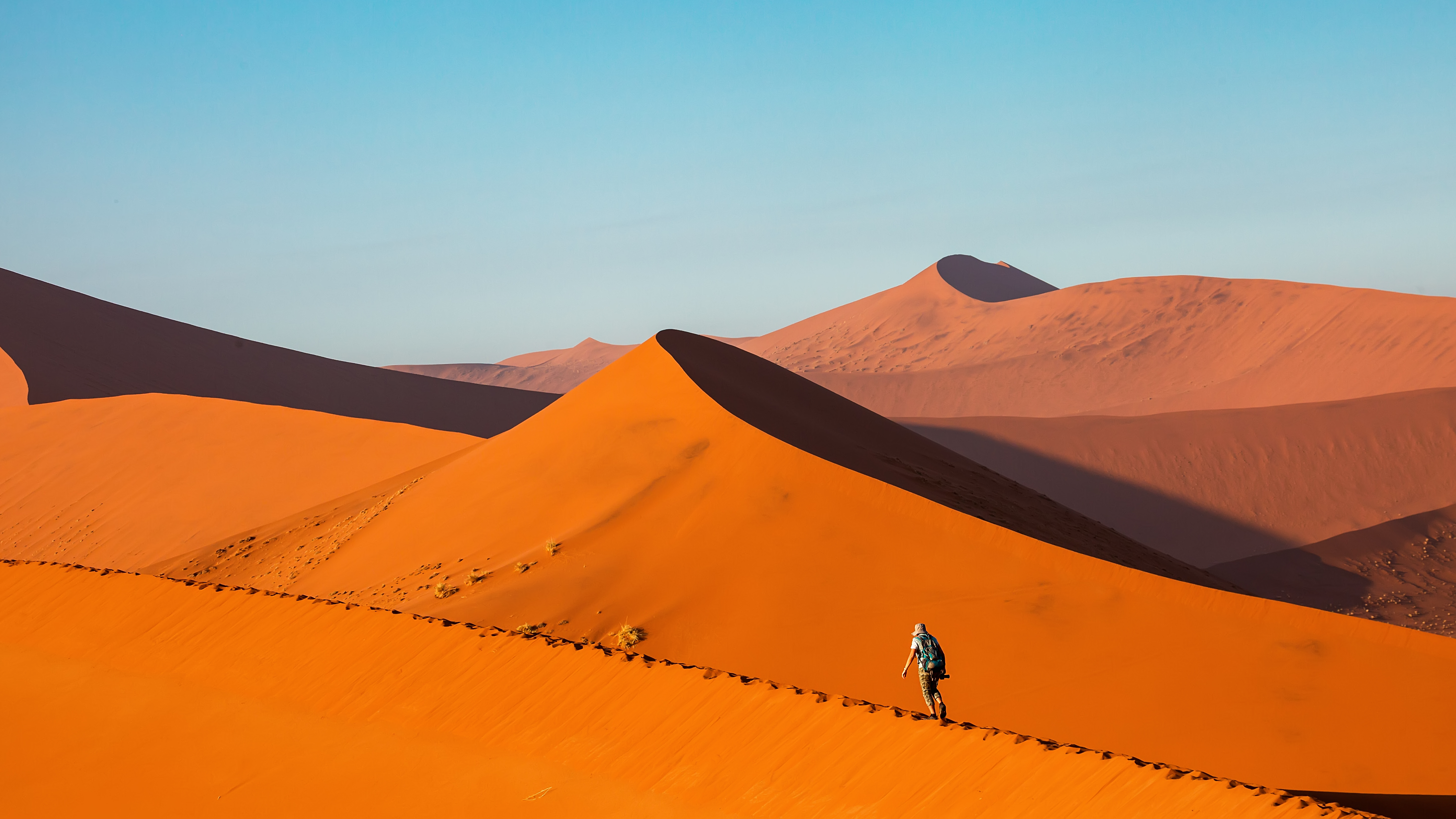 Hiker walks along a sharp dune ridge at Sossusvlei, with layered orange dunes and deep shadow folds behind