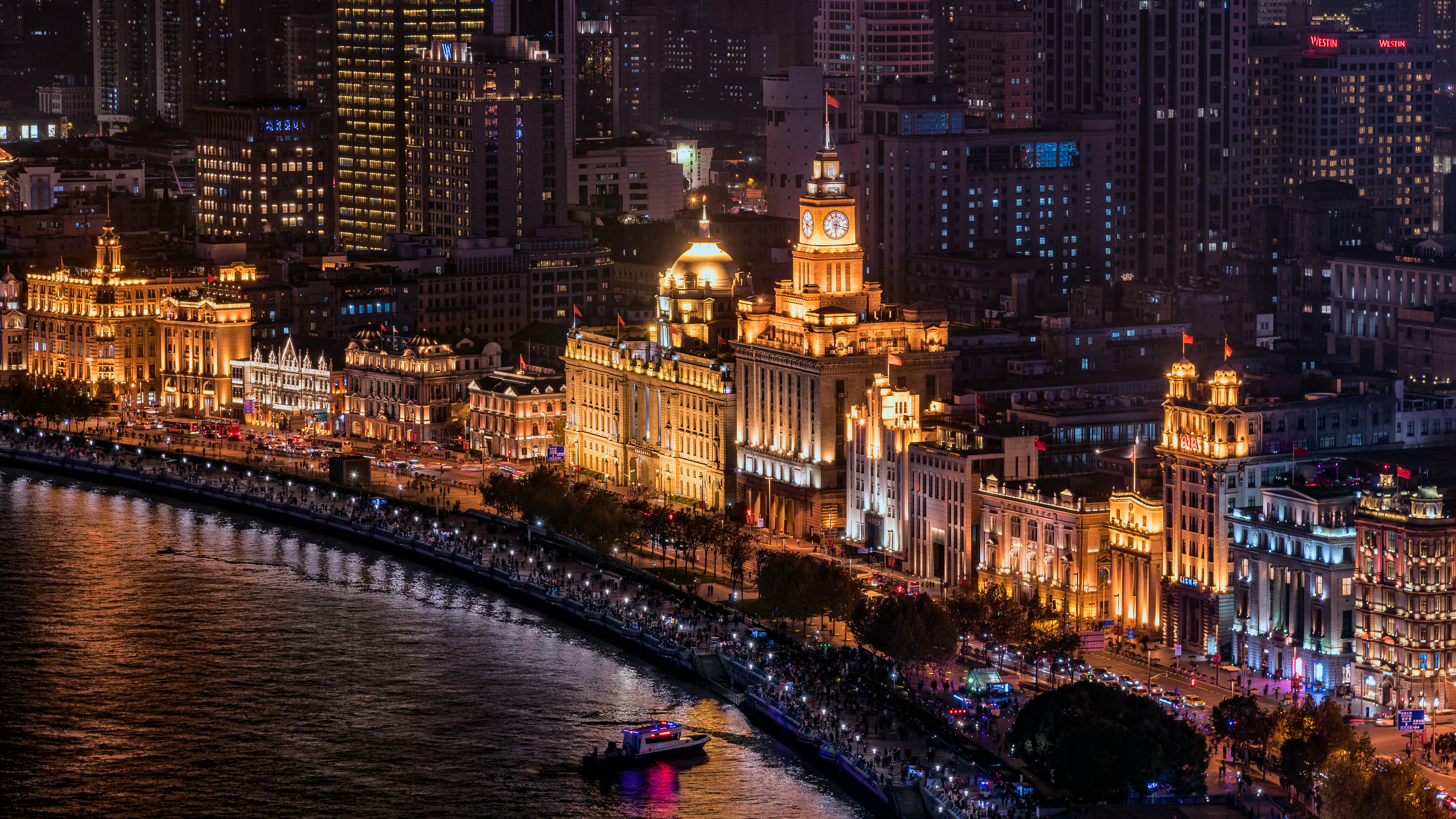 Illuminated colonial-era buildings along the Bund in Shanghai at night, with a boat passing on the river and crowds along the promenade