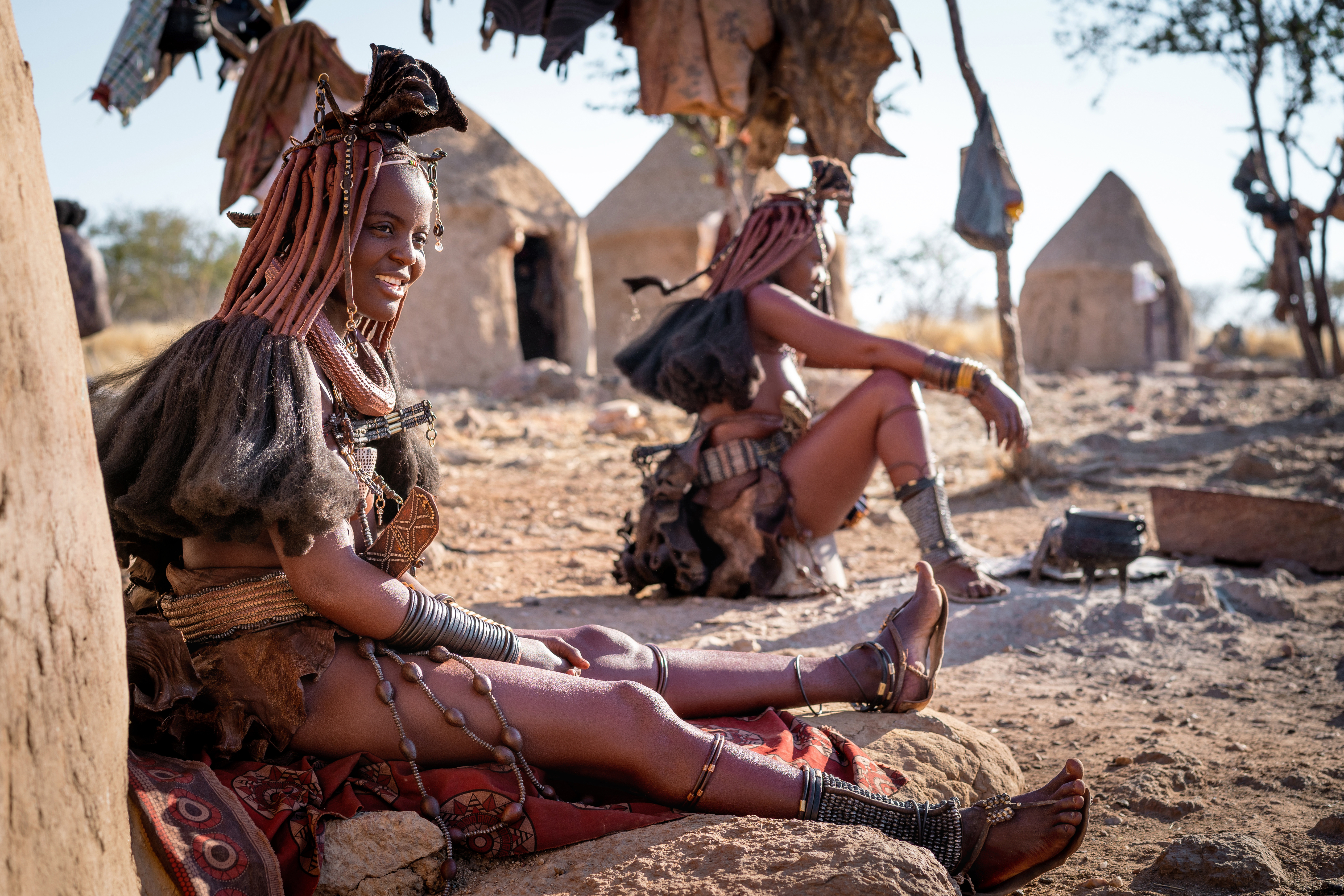 Himba women with traditional ochre-tinted braided hair and beaded jewelry sit outside conical huts in a Kaokoland village in northern Namibia