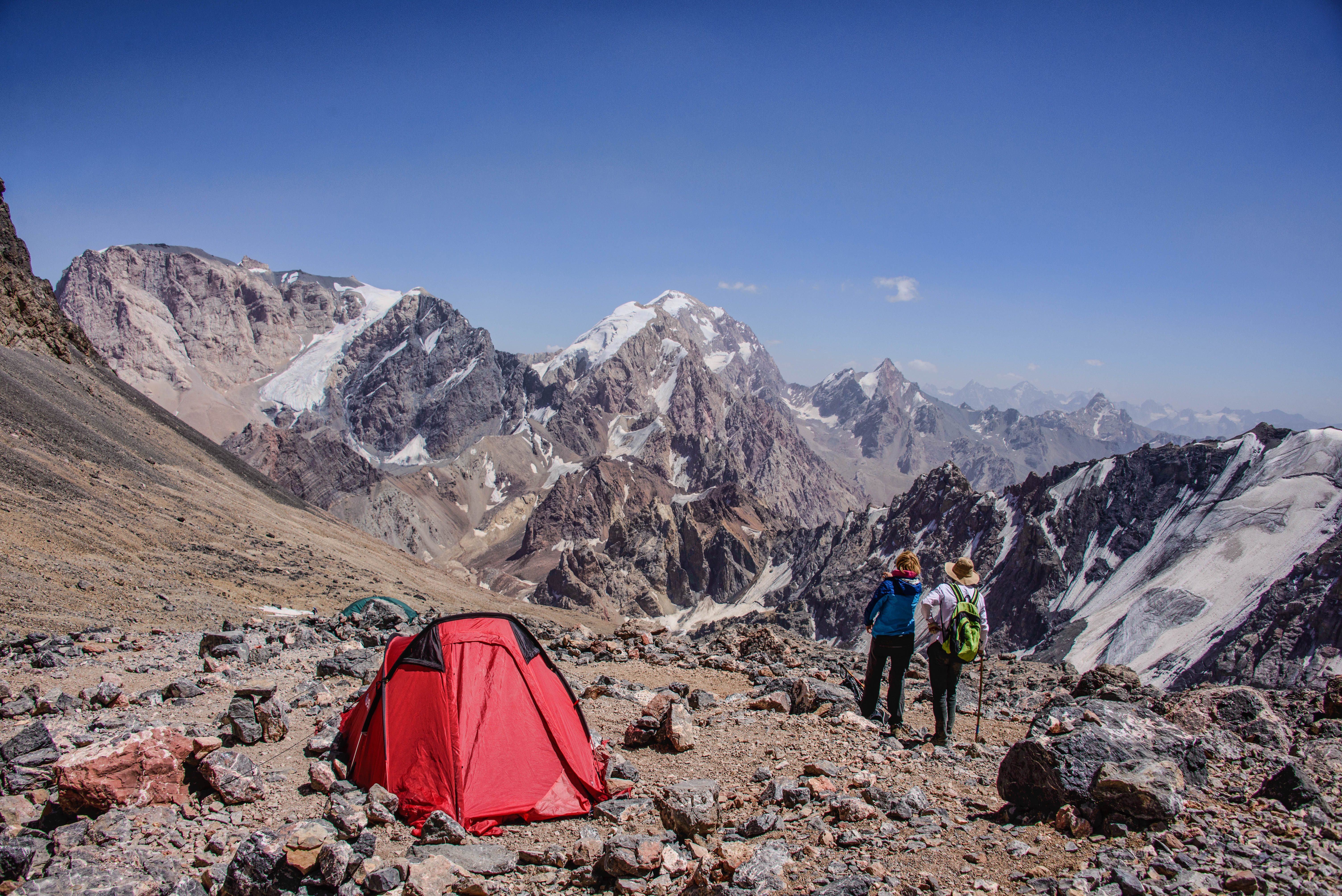 Two hikers pause near a red tent on a rocky slope, overlooking snow-streaked peaks along the Chimtarga Pass route, Tajikistan