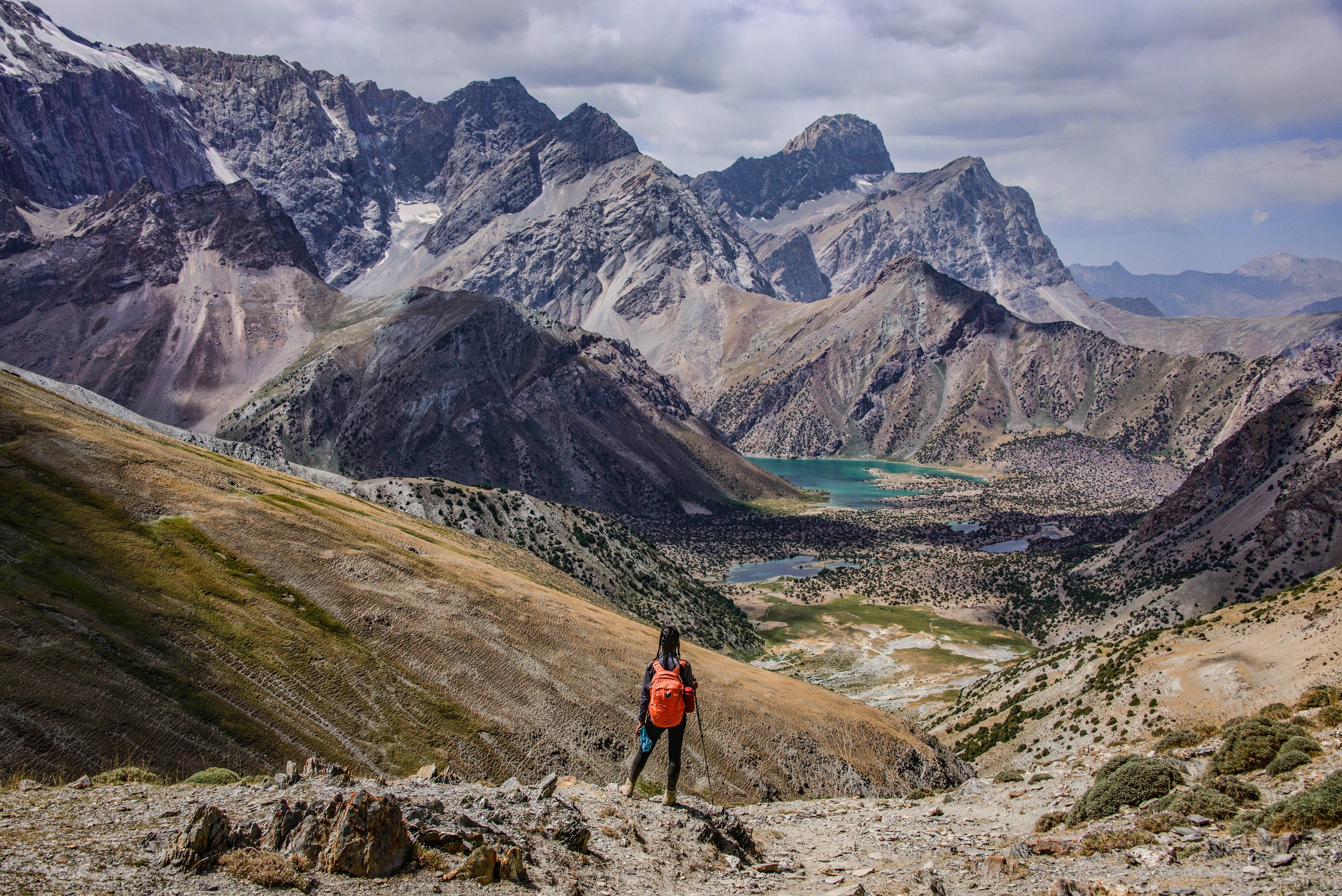 Hiker with an orange backpack descends a rocky slope toward a turquoise lake in the Fann Mountains, Tajikistan, snow-streaked peaks beyond