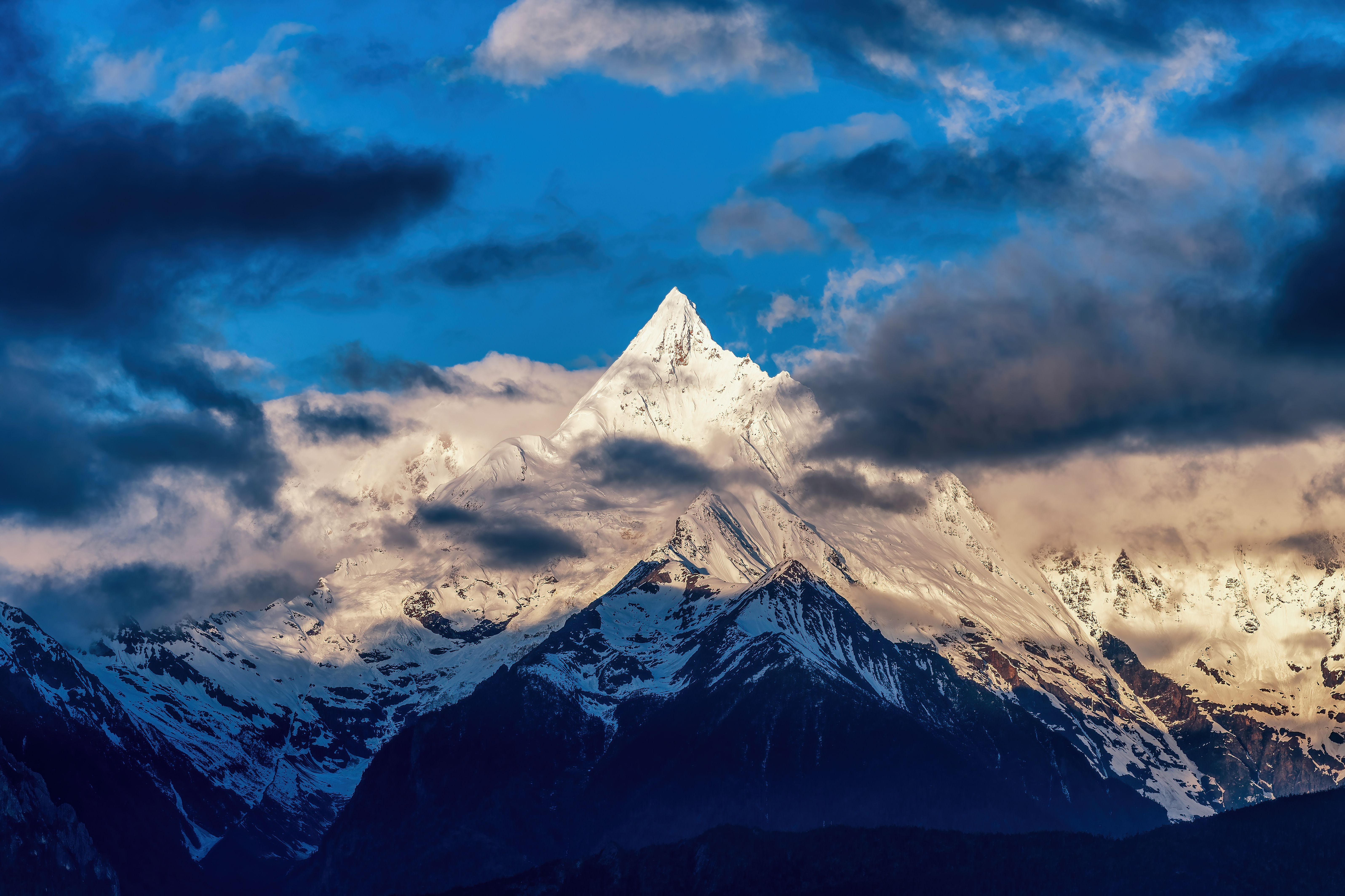 Snow-capped peak of Meili (Kawakarpo) mountain lit golden at sunrise, surrounded by drifting clouds in Yunnan, China