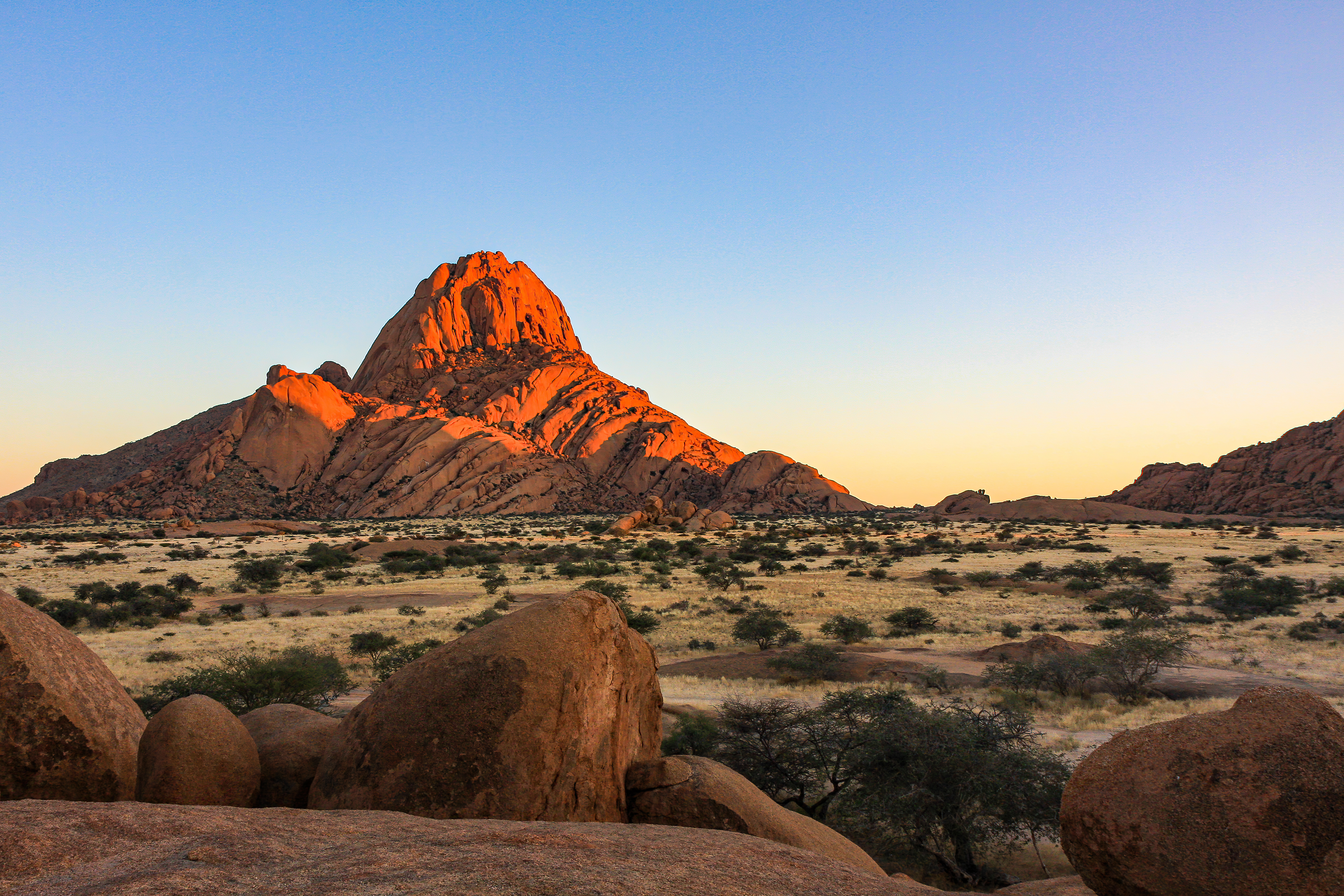 Spitzkoppe's granite peak glows orange at sunset above boulders and dry scrubland in Namibia
