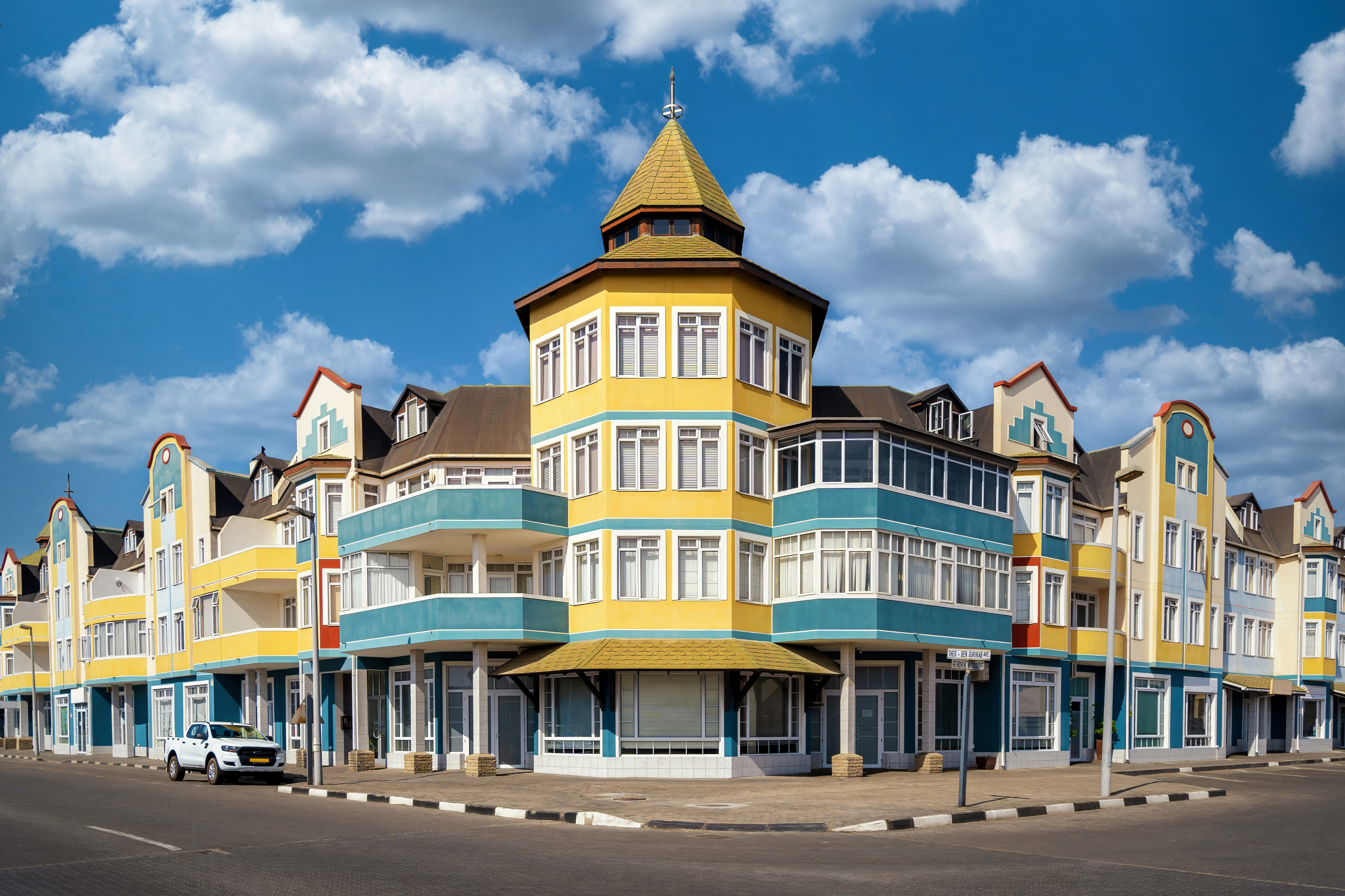 Turreted corner building in Swakopmund painted yellow and teal with white-trimmed windows, under a partly cloudy blue sky