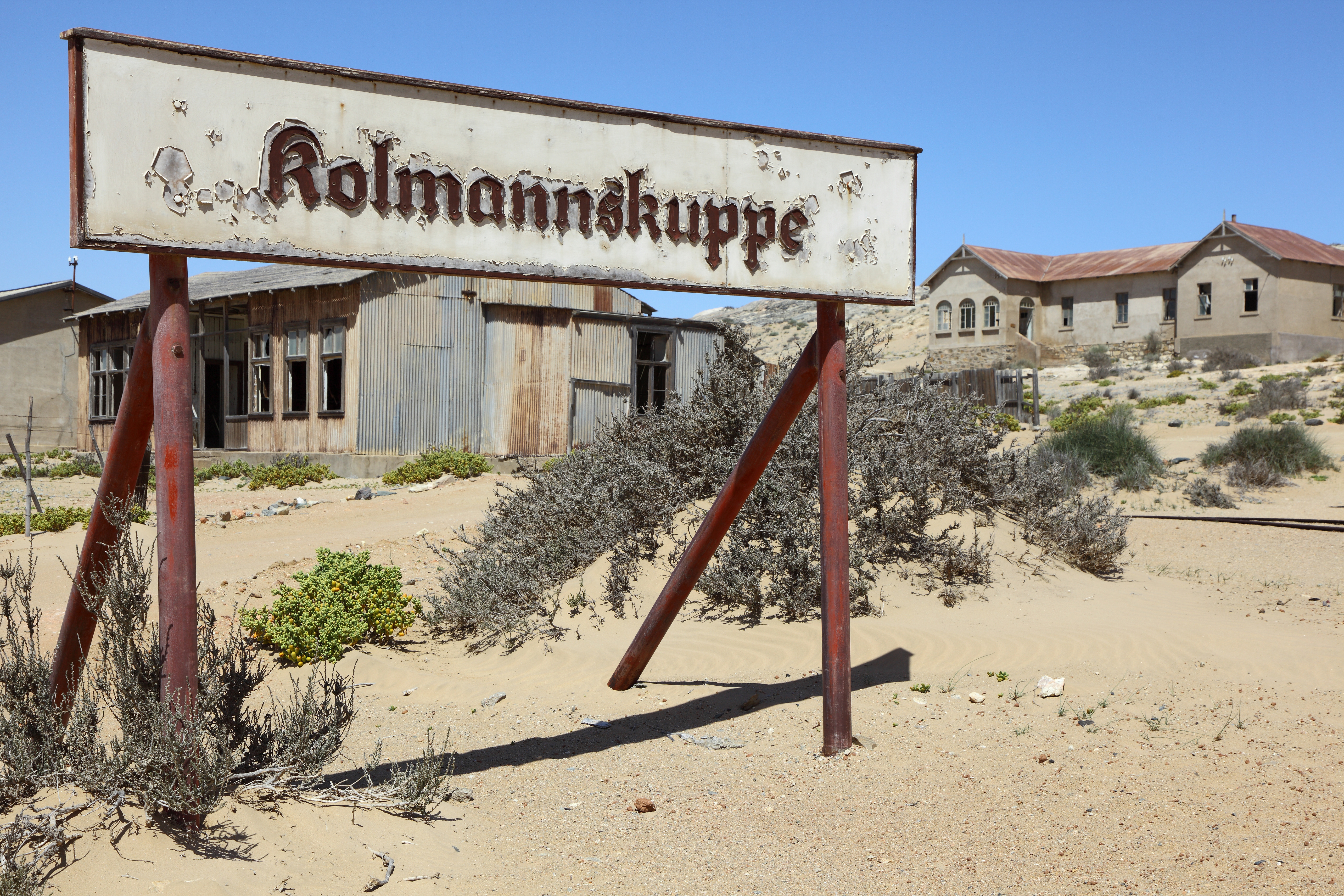 Weathered sign reading Kolmannskuppe stands on rusted posts amid desert sand, with abandoned corrugated buildings in the background