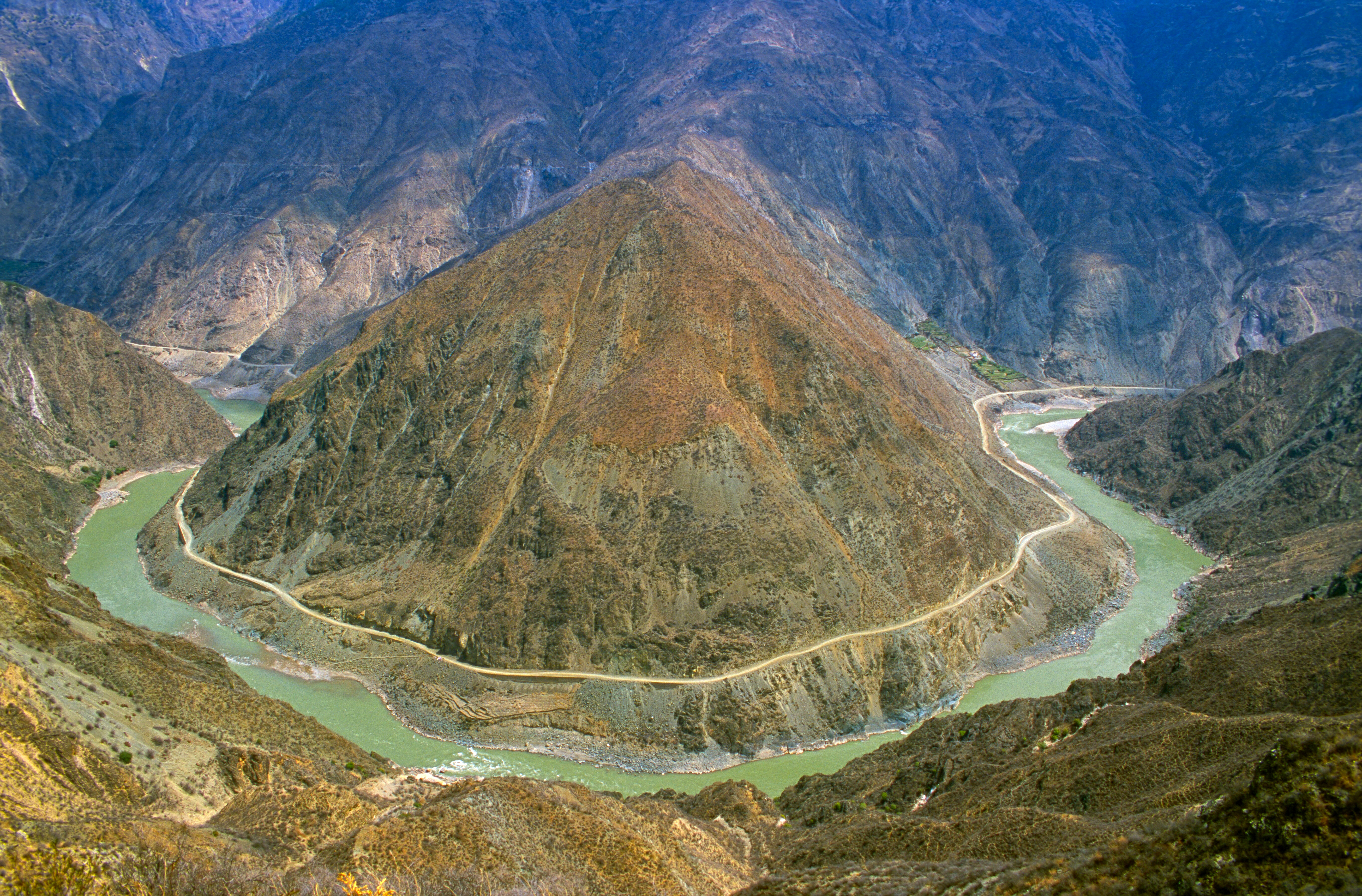 A river makes a sharp horseshoe bend around a pyramid-shaped mountain, with a road tracing the curve of the water at the First Bend of the Yangtze, Yunnan
