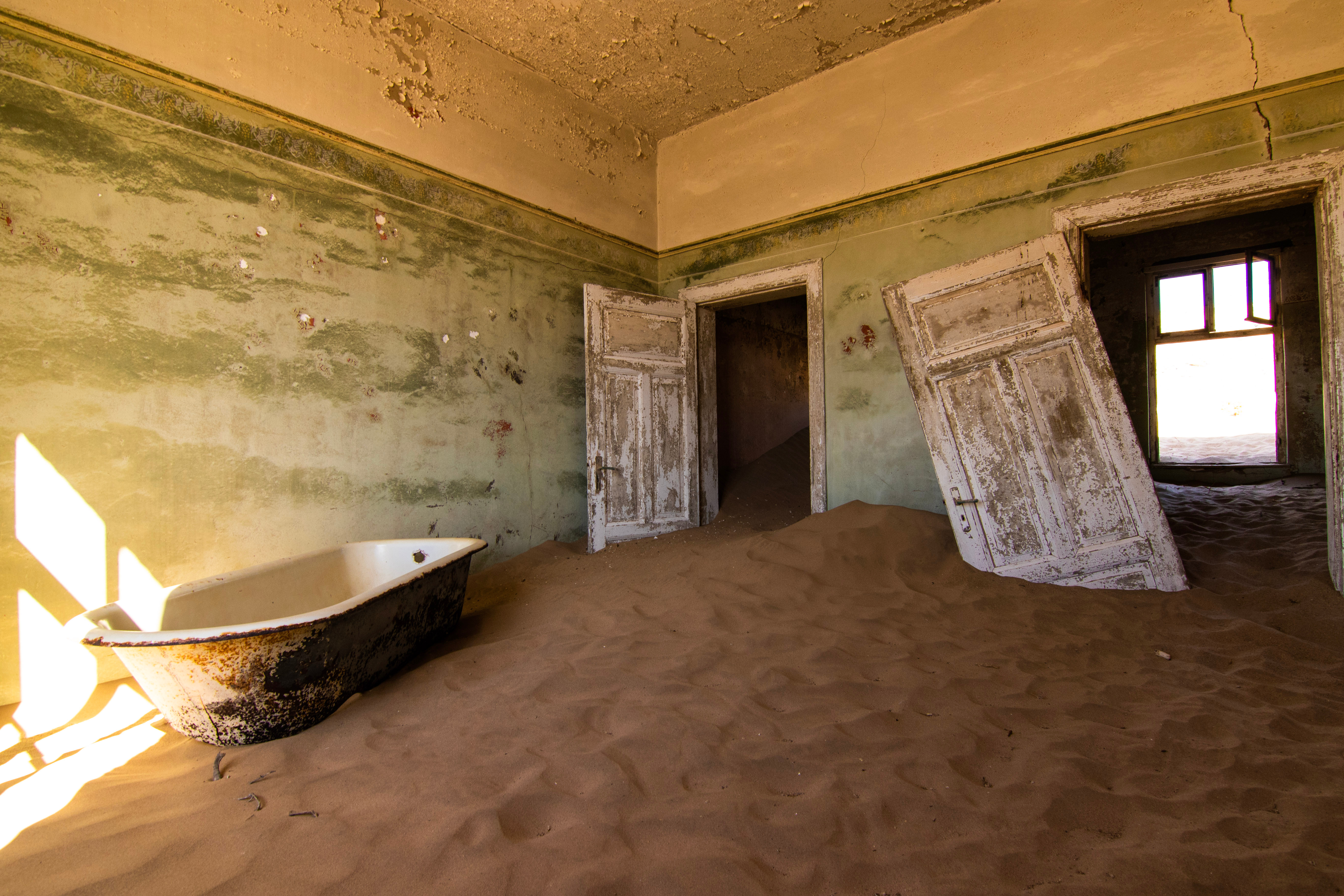 Sand drifts across the floor of an abandoned room at Kolmanskop, Namibia, with a rusted bathtub and peeling doors