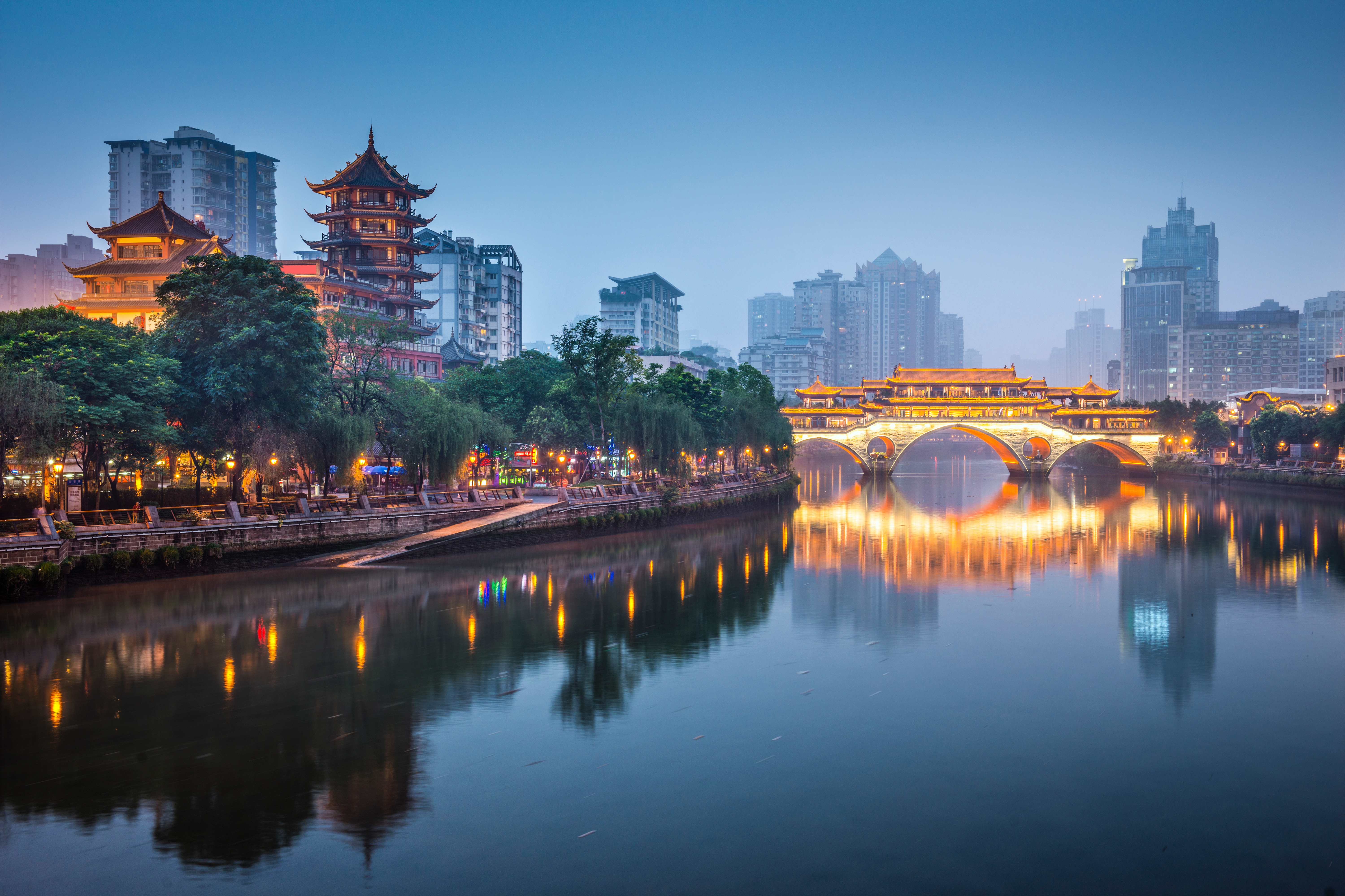 Illuminated Anshun Bridge arches over the Jin River at dusk in Chengdu, Sichuan, with a lit pagoda and city skyline reflected in the water.