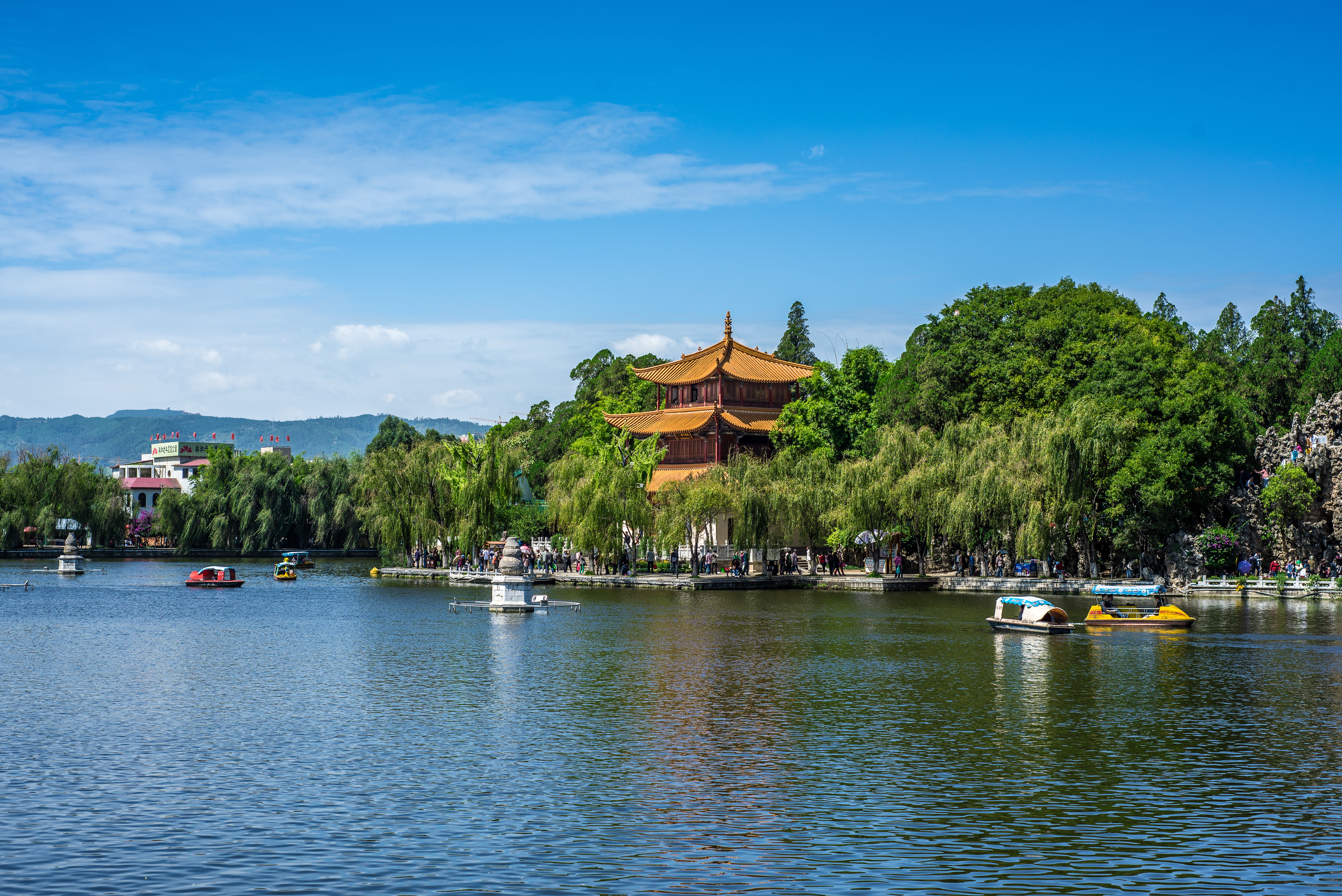Traditional Chinese pagoda with orange tiered roof rises above willow trees beside a lake in Kunming, with pedal boats on the water