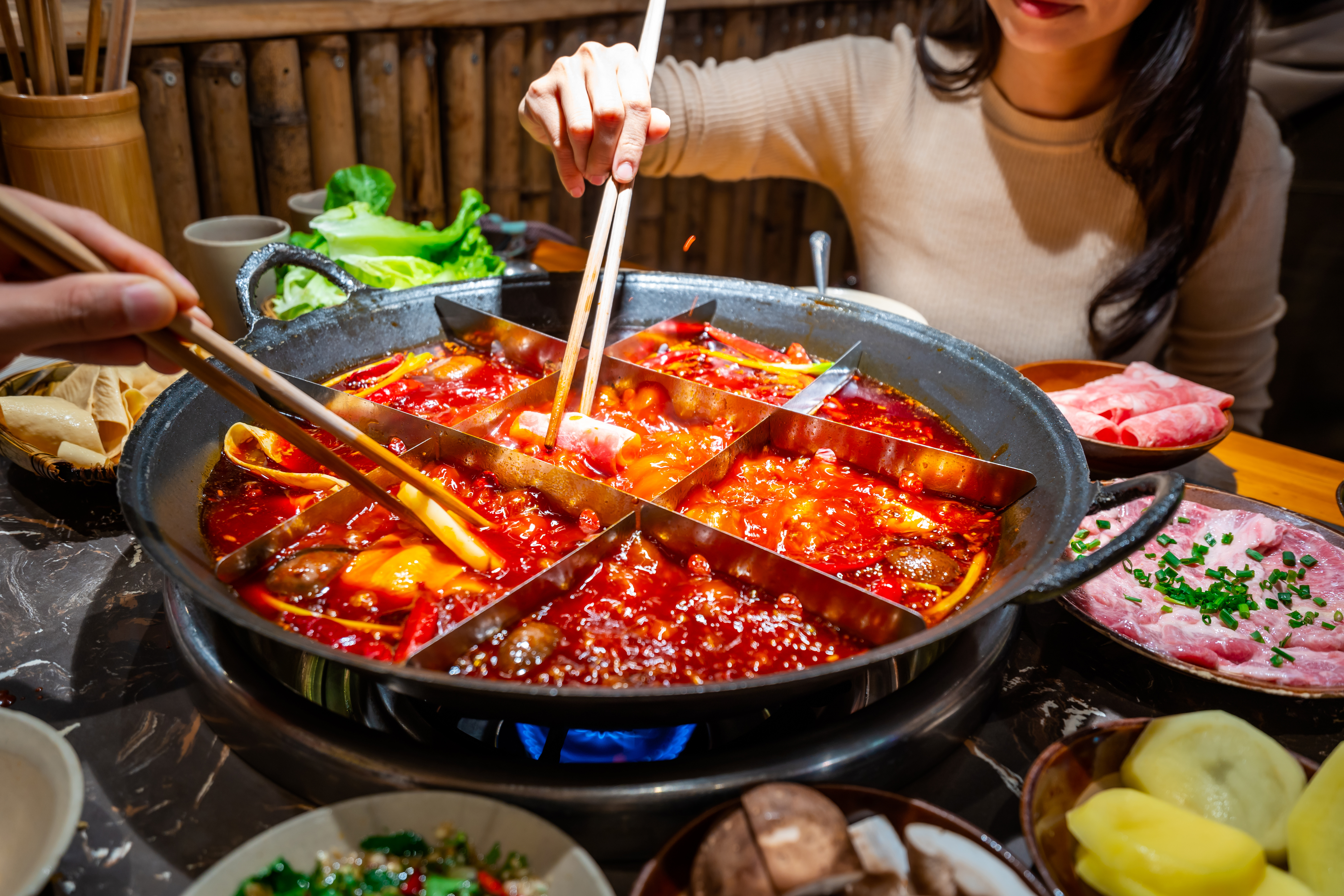 Divided hot pot with a fiery mala broth on one side, diners using chopsticks to dip meat and vegetables at the table