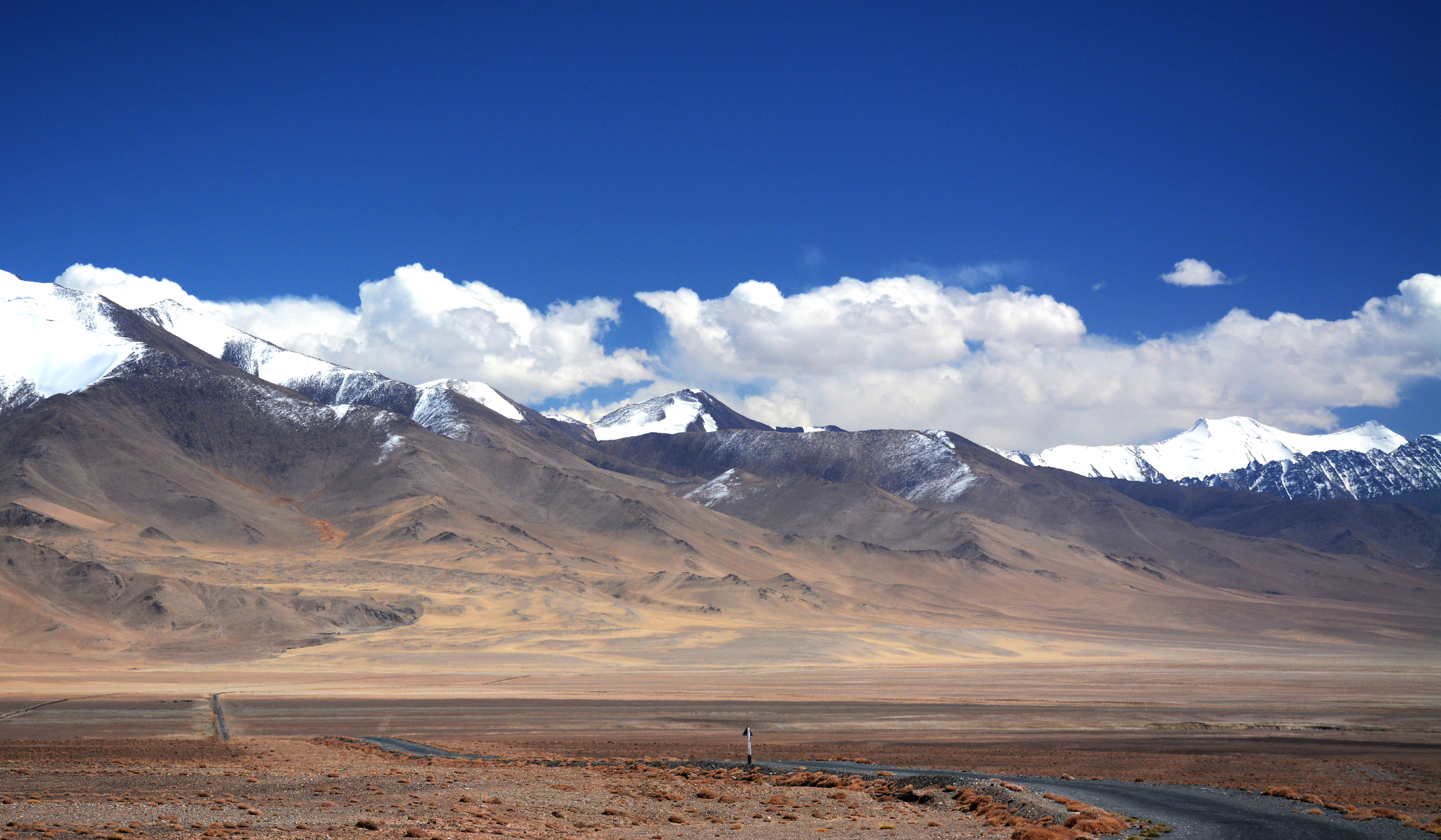 Snow-capped Pamir peaks rise above a broad arid plateau along the Pamir Highway near Karakul Lake, Tajikistan, with clouds hanging low over the ridgeline