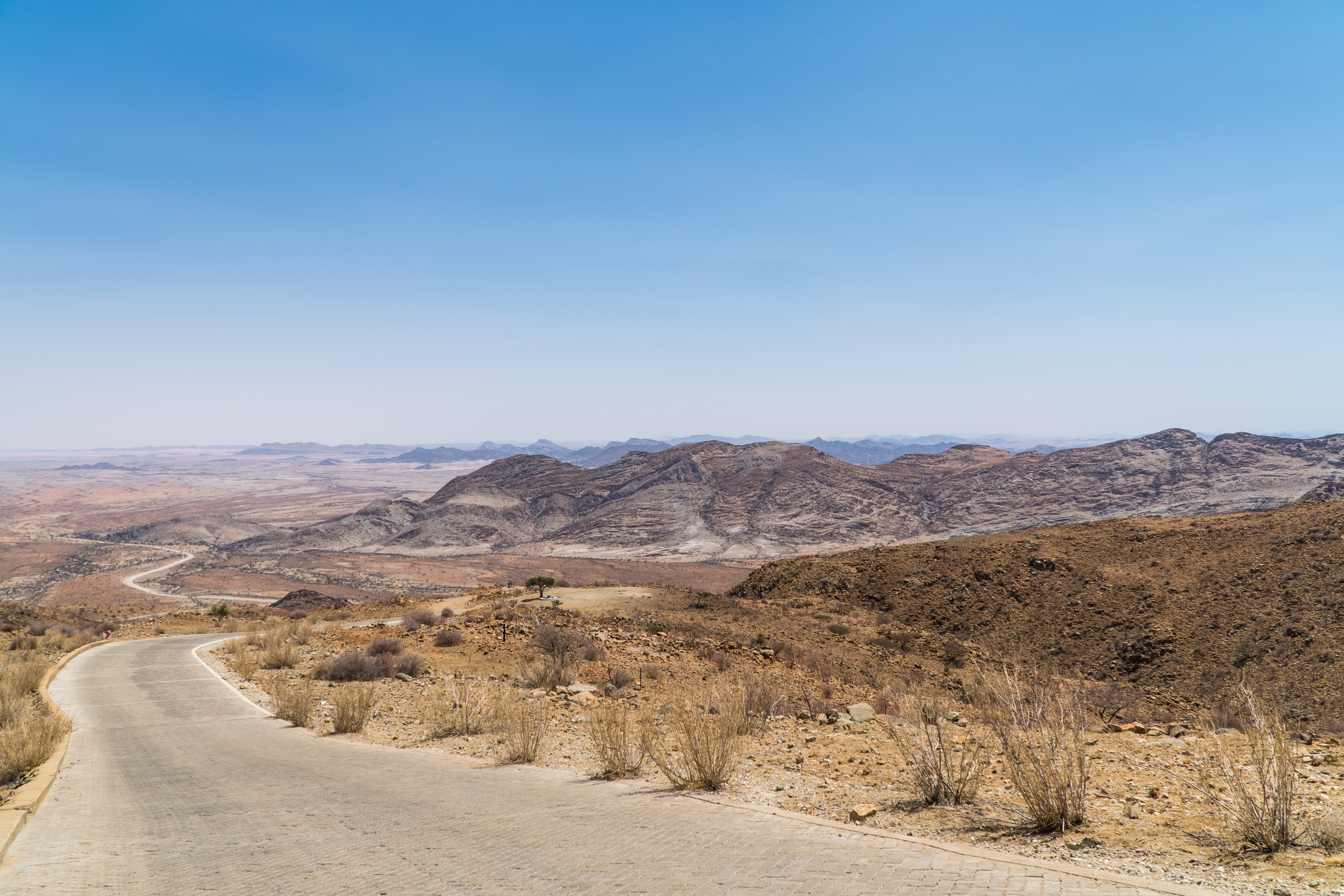 Paved road winds down a mountain pass toward a vast arid plain, with layered rocky peaks stretching to the horizon under a hazy sky