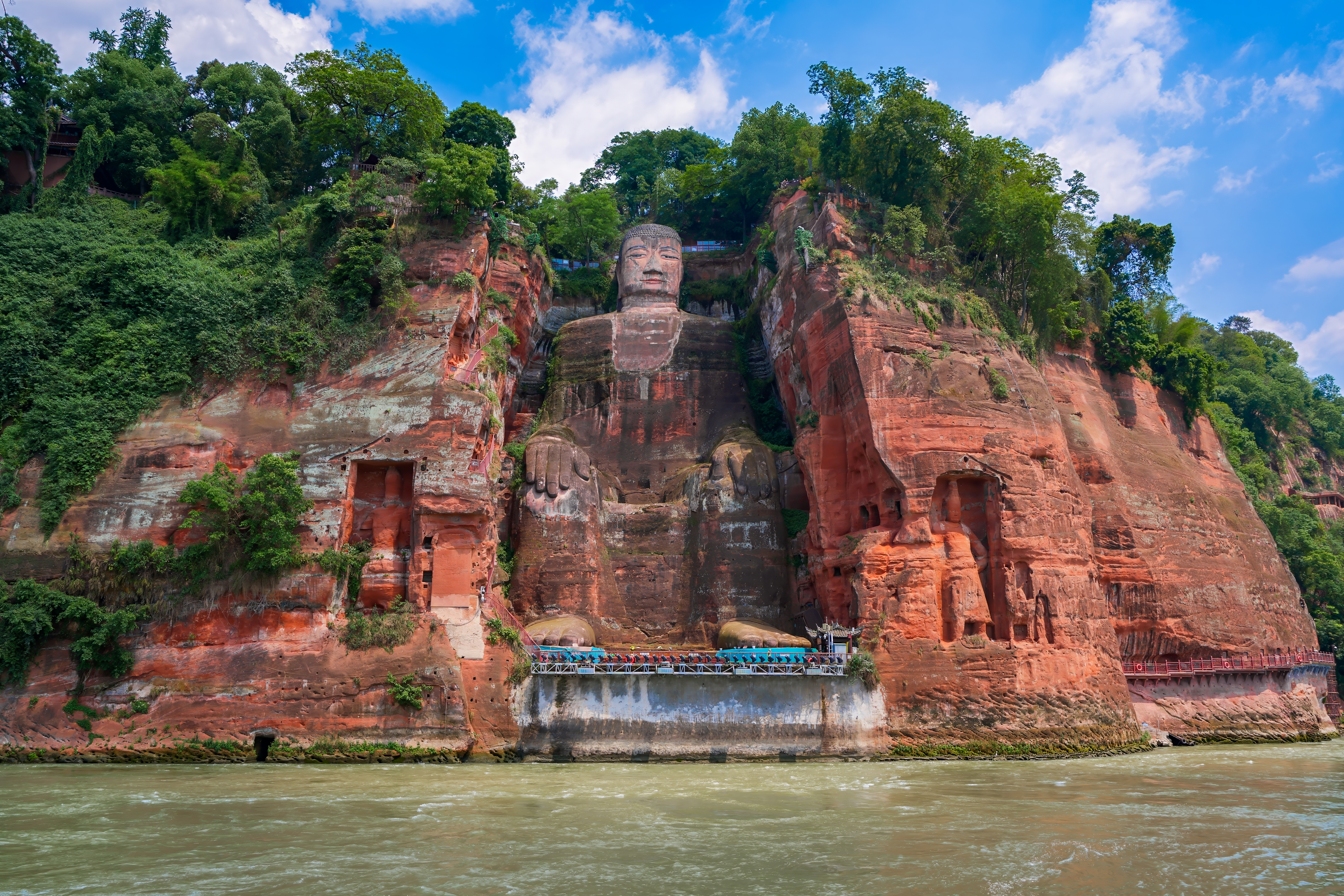 The Leshan Giant Buddha carved into a red sandstone cliff above the river, seen from the water with hands resting on its knees