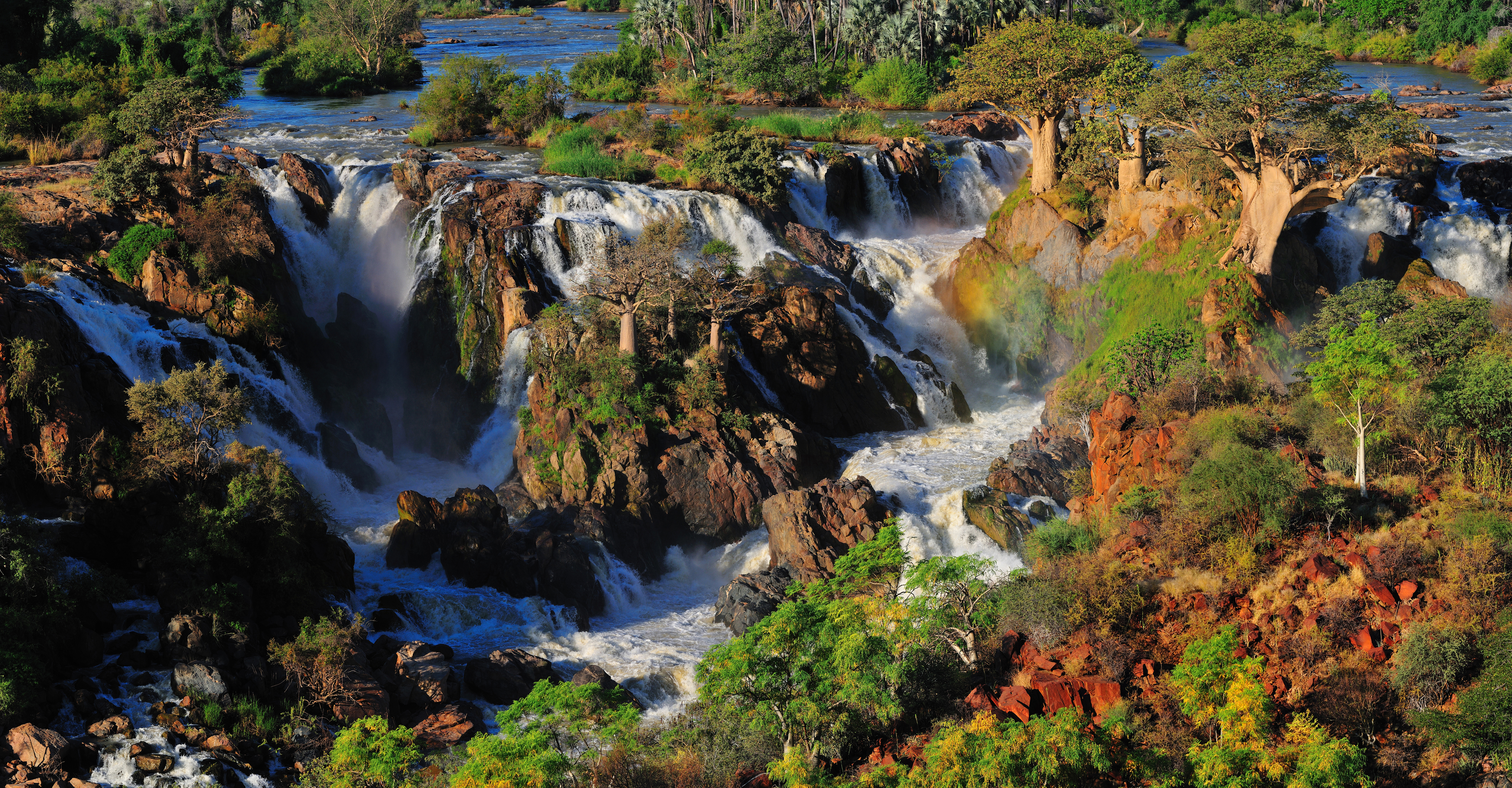 Multiple cascades of the Epupa waterfalls drop over dark rock, with baobab trees on the islands and a faint rainbow in the spray