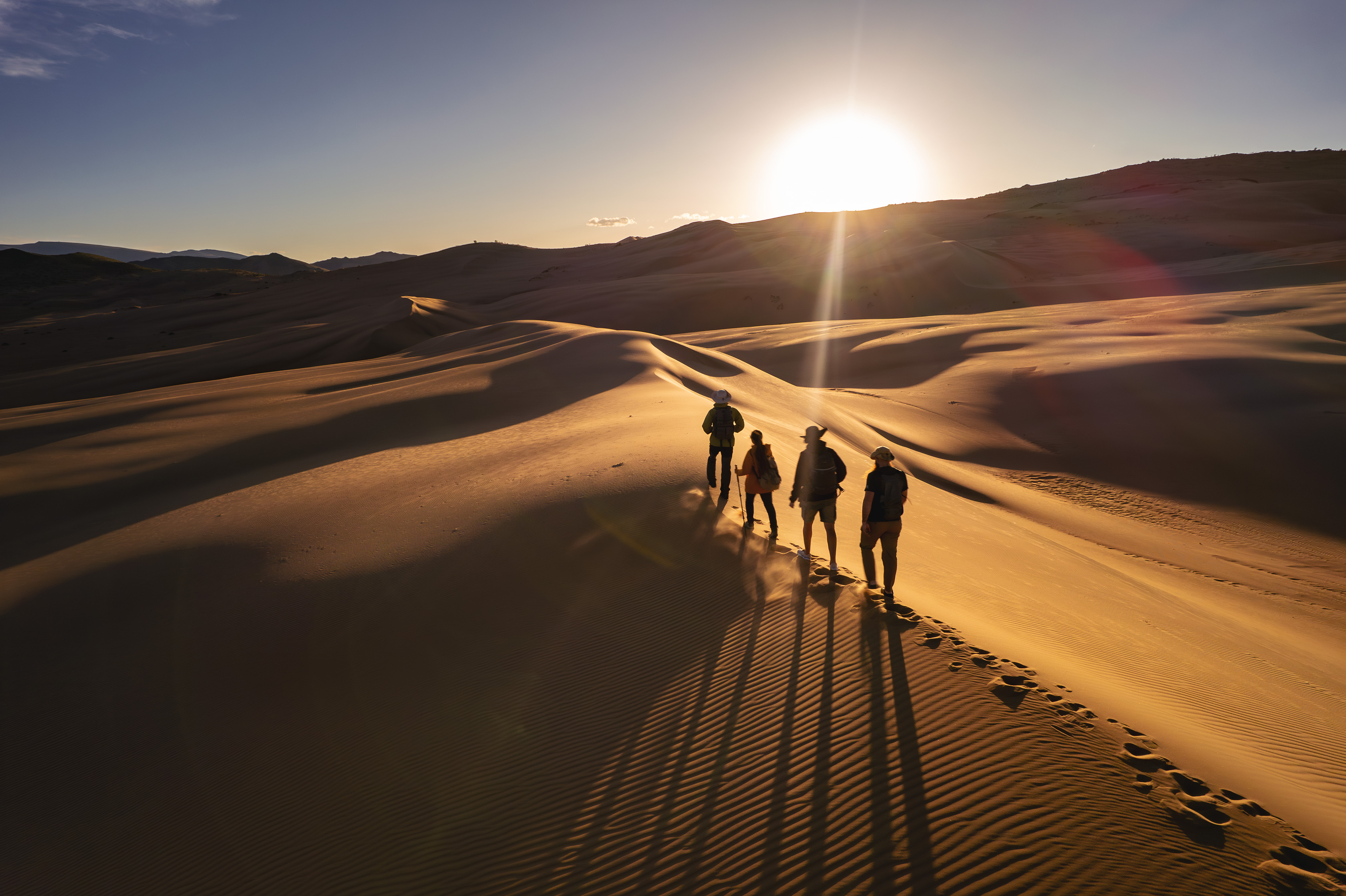 Four hikers with backpacks walk along a sand dune ridge in the Gobi Desert, Mongolia, casting long shadows at sunset