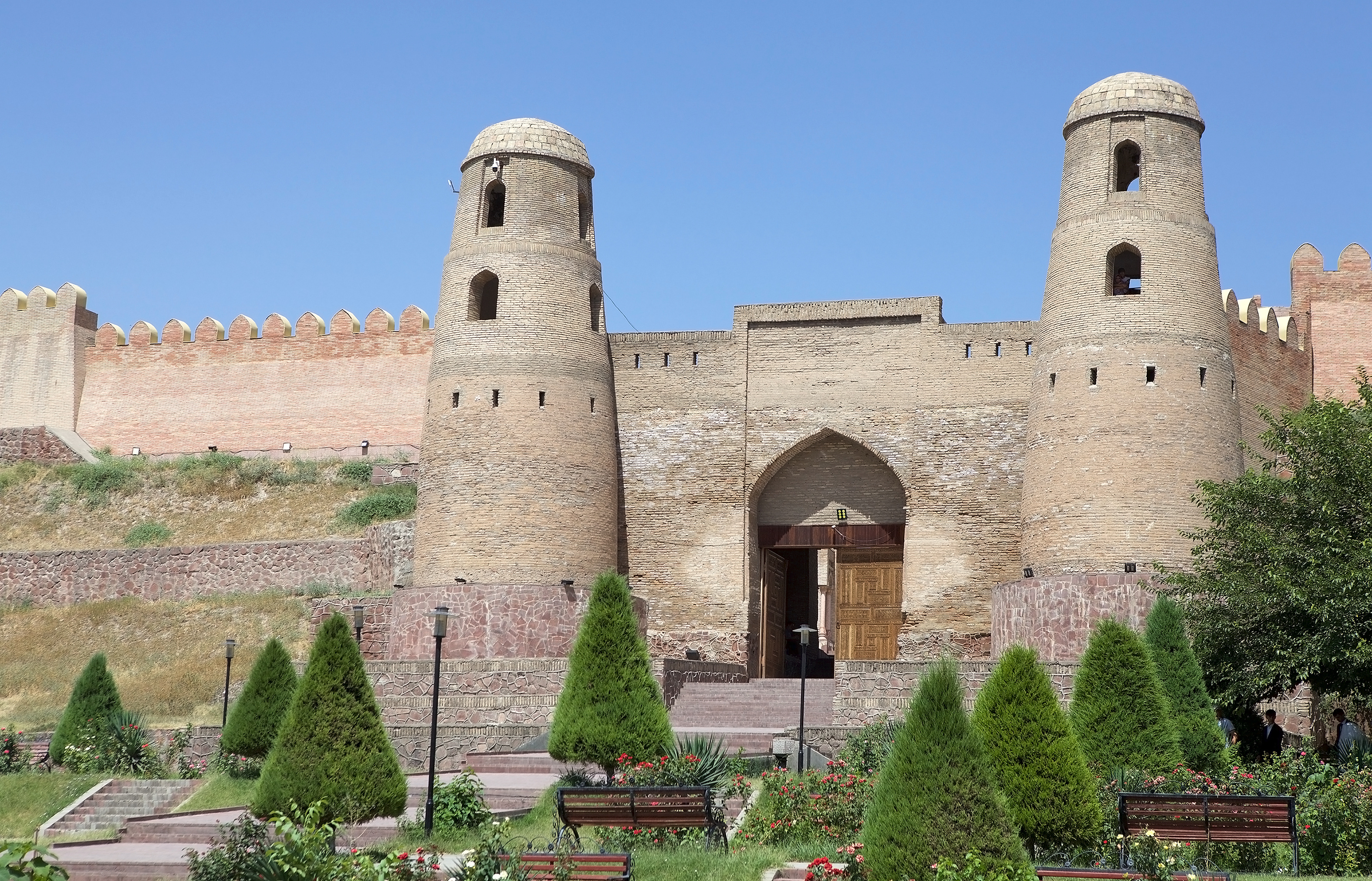 Twin round towers flank the reconstructed gateway of Hissar Fortress near Dushanbe, with a landscaped garden of conifers and roses below