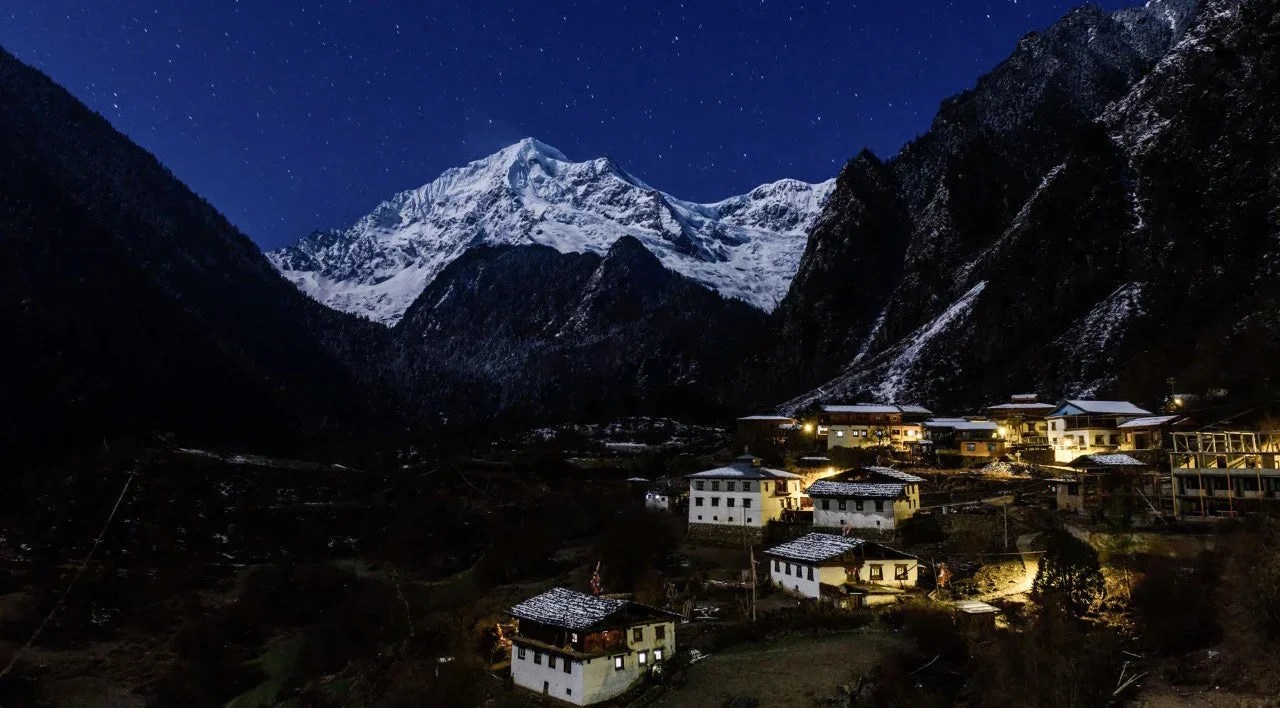 Snow-capped peak glows under starlight above a lit village of stone and timber houses near Deqin, Yunnan
