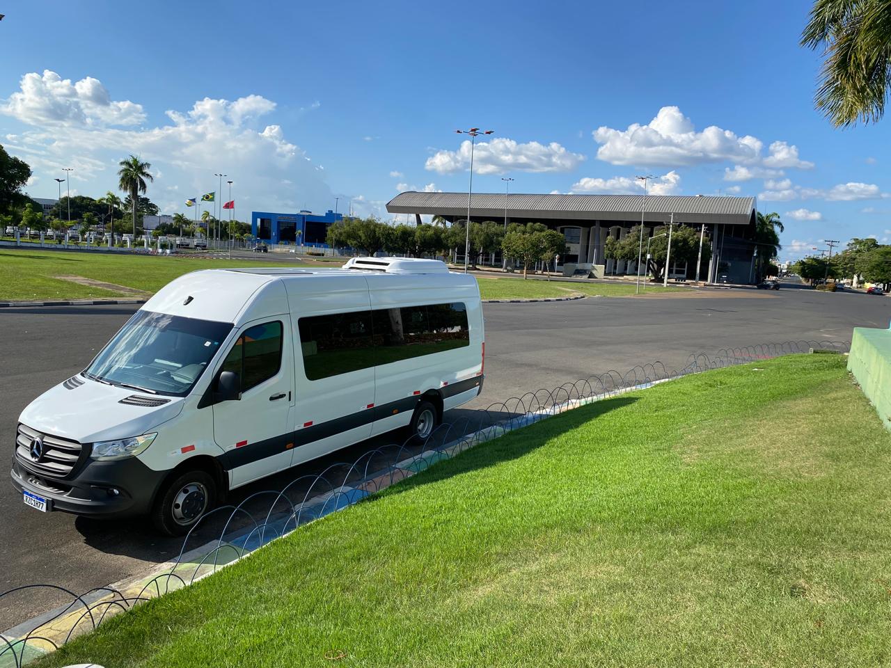 White passenger van parked roadside near a flag plaza and a large open-sided building under a partly cloudy sky