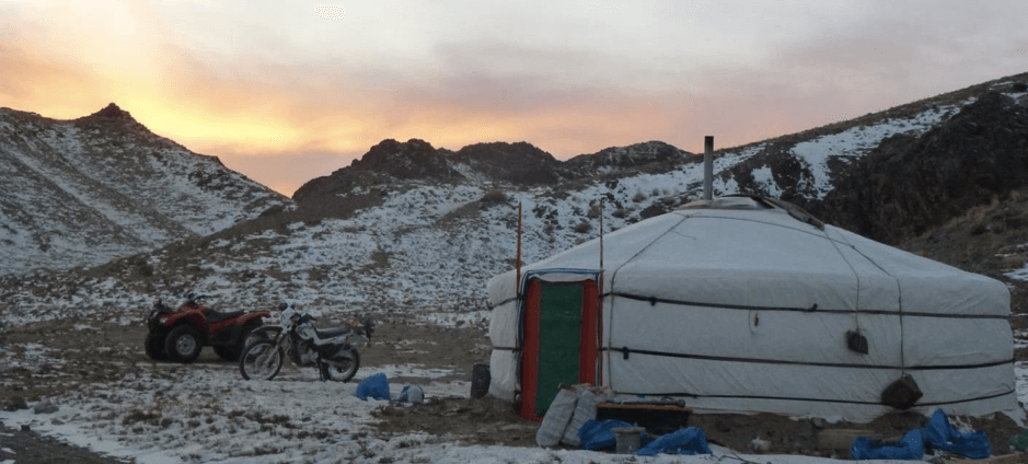 A traditional felt yurt with stovepipe stands beside parked motorbikes on snowy ground, with snow-dusted mountains under a dawn sky in Kyrgyzstan
