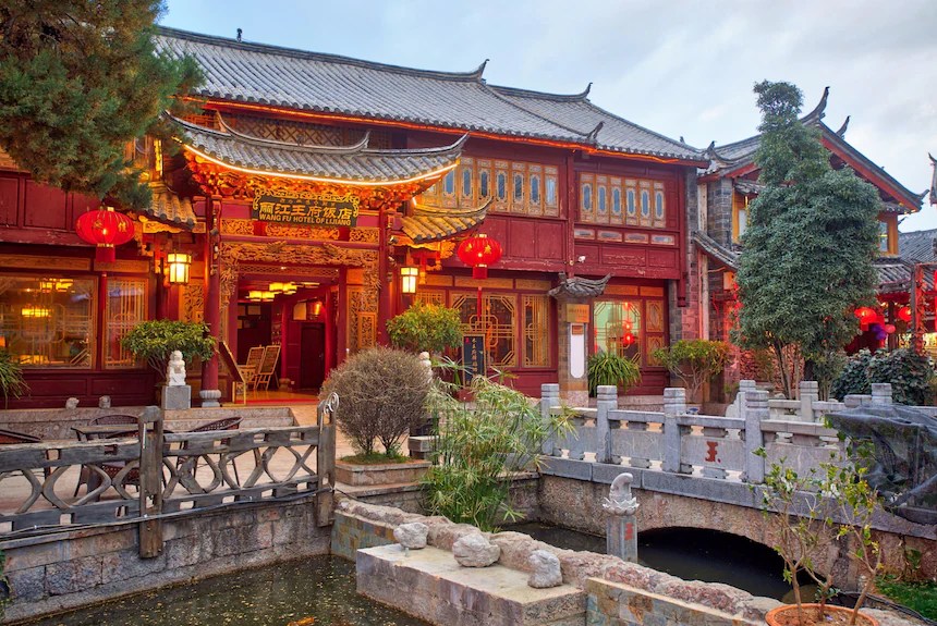 Red and gold facade of the Wangfu Hotel in Lijiang at dusk, with lanterns lit and a stone bridge over a small canal in front