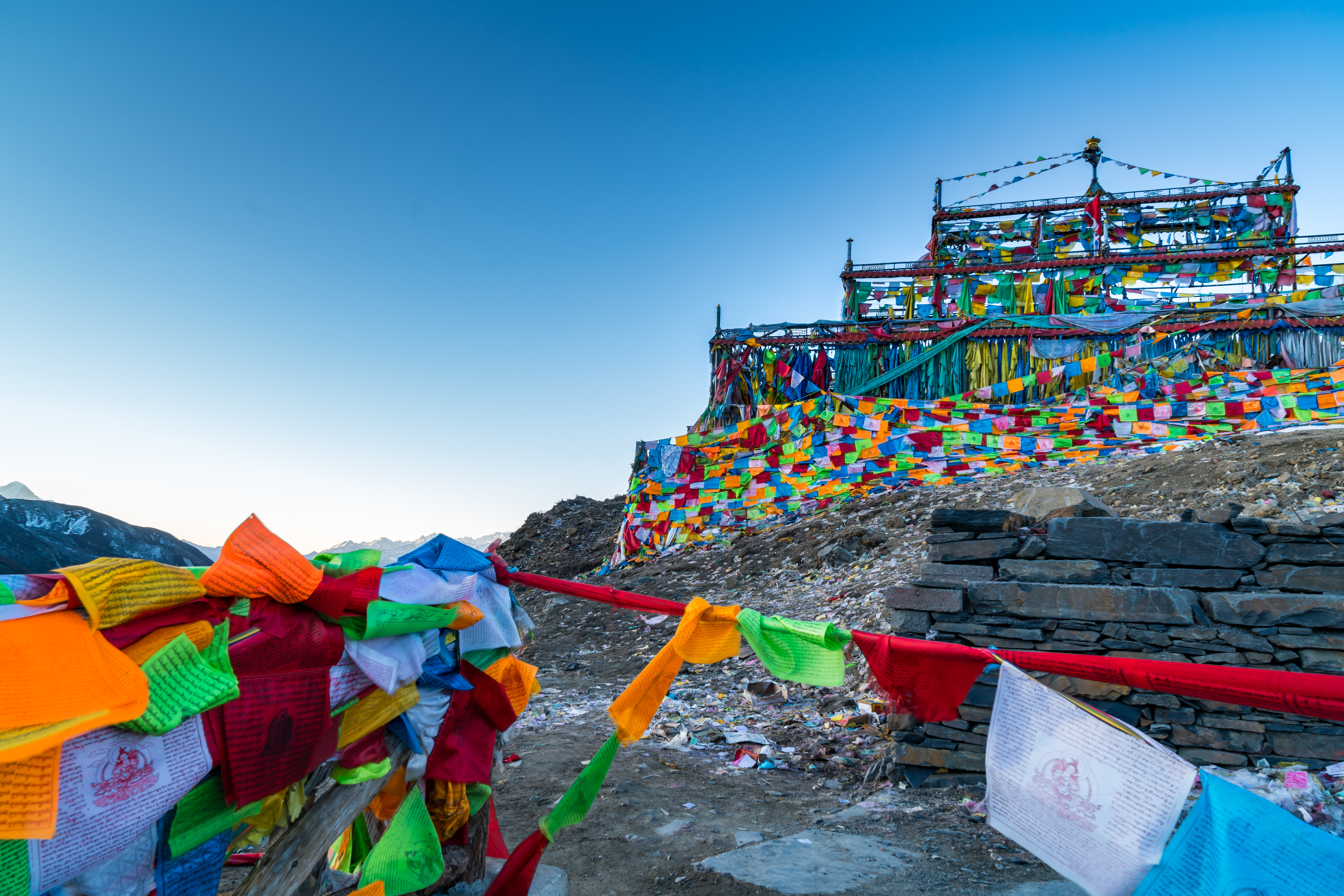 Thousands of multicolored prayer flags cover a hillside shrine structure at Zheduo Shan Pass, with clear blue sky and distant mountains