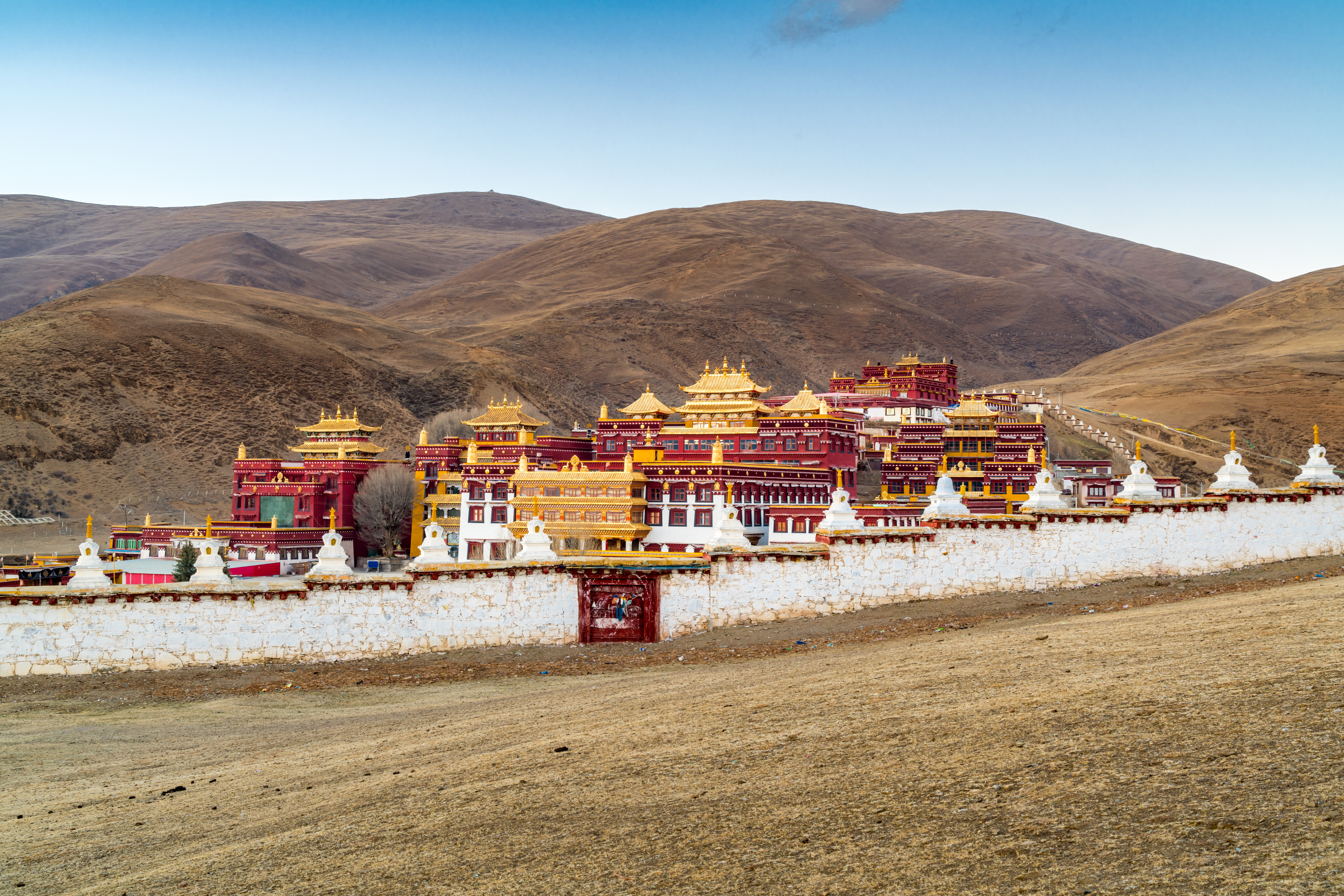 Red and gold Tibetan monastery buildings with white stupas behind a whitewashed wall, set against bare brown hills in Litang, Sichuan