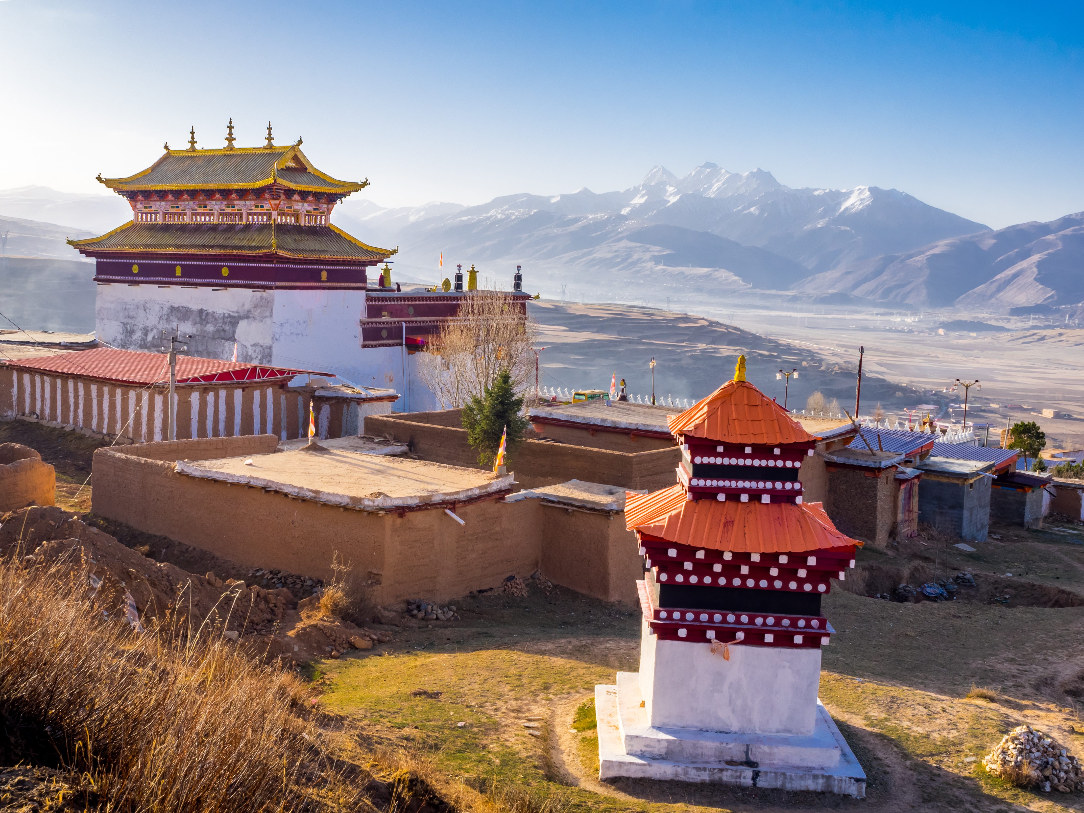 Red and white Tibetan monastery with golden roof ornaments in Dege, with a painted stupa in the foreground and snow peaks beyond