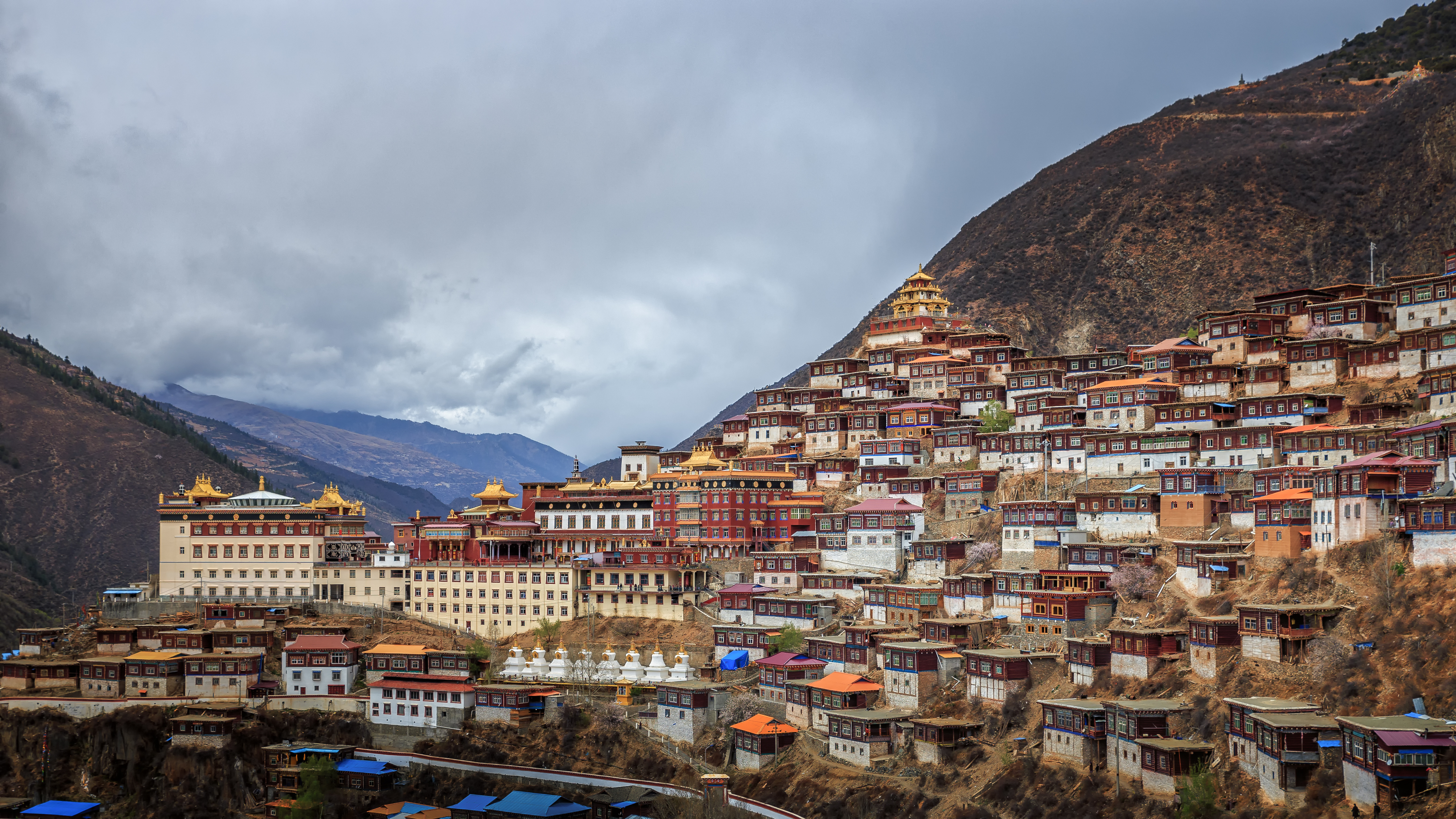 Maroon and white Tibetan monastery buildings of Gengqing Monastery cover a steep hillside above Dege, with mountains behind under clouds