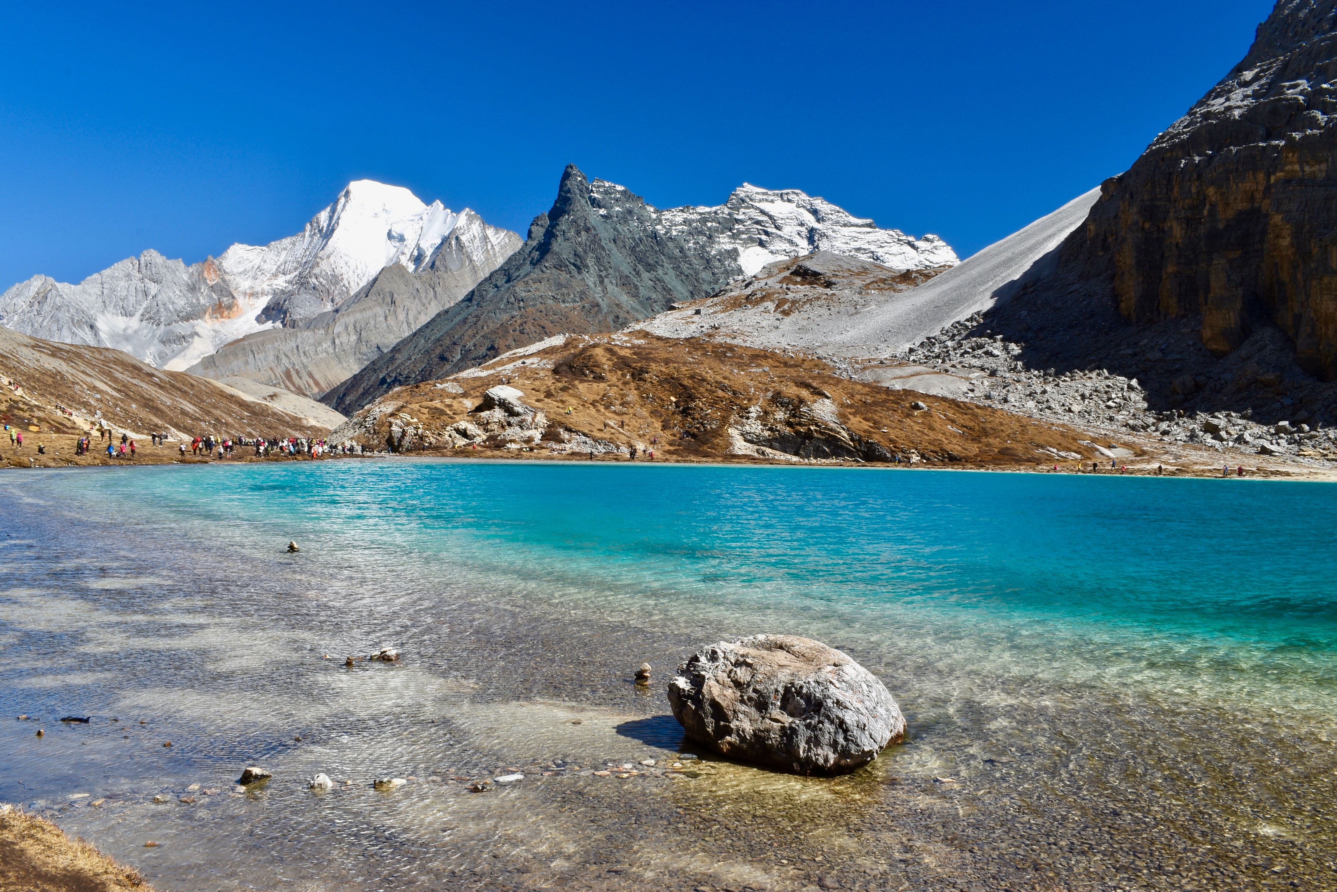 Turquoise Milk Lake beneath snow capped peaks at Yading Nature Reserve, with visitors gathered along its shore