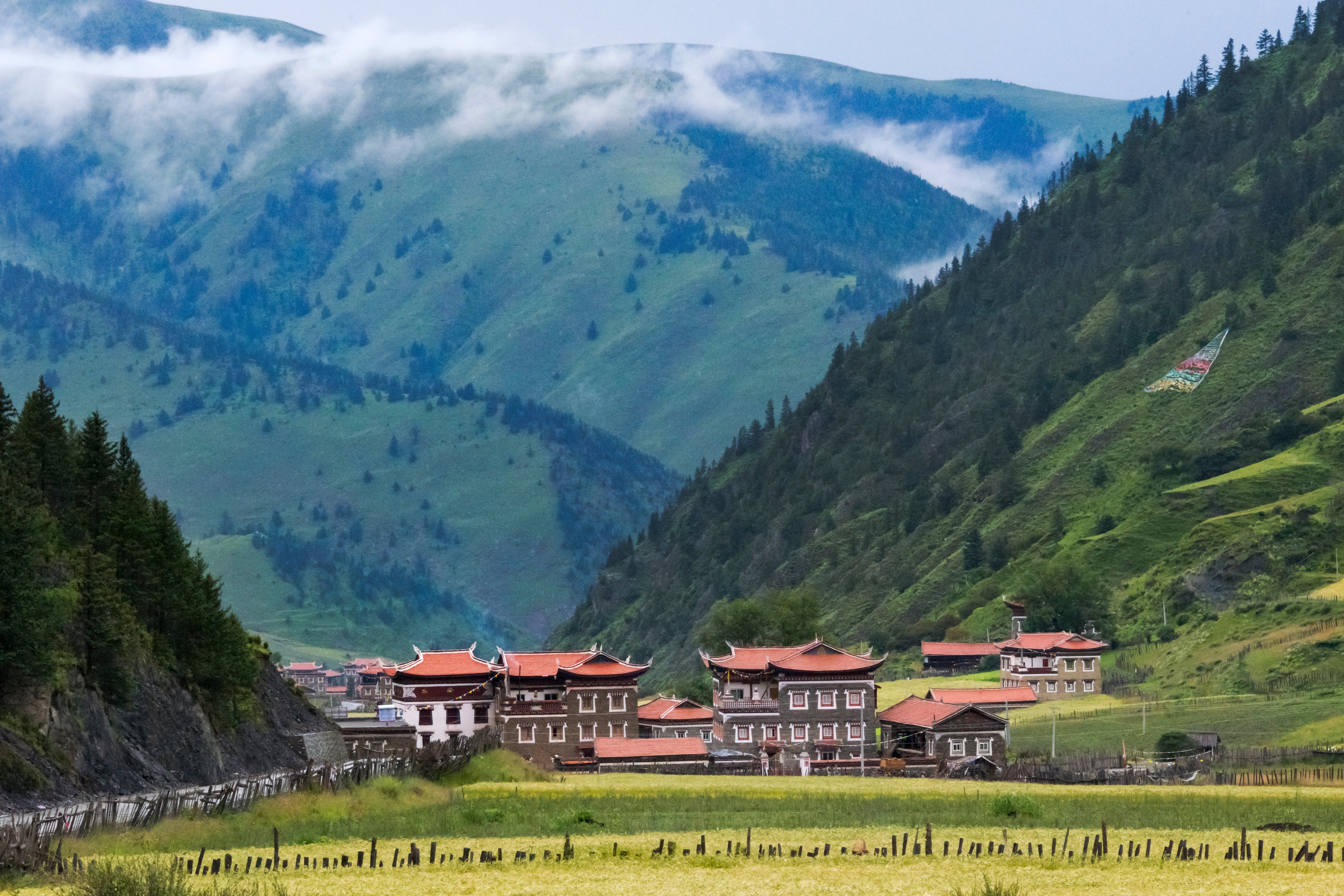 Tibetan village with ochre and white houses and orange rooftops set in a grassland valley near Xinduqiao, forested mountains rising behind under drifting clouds
