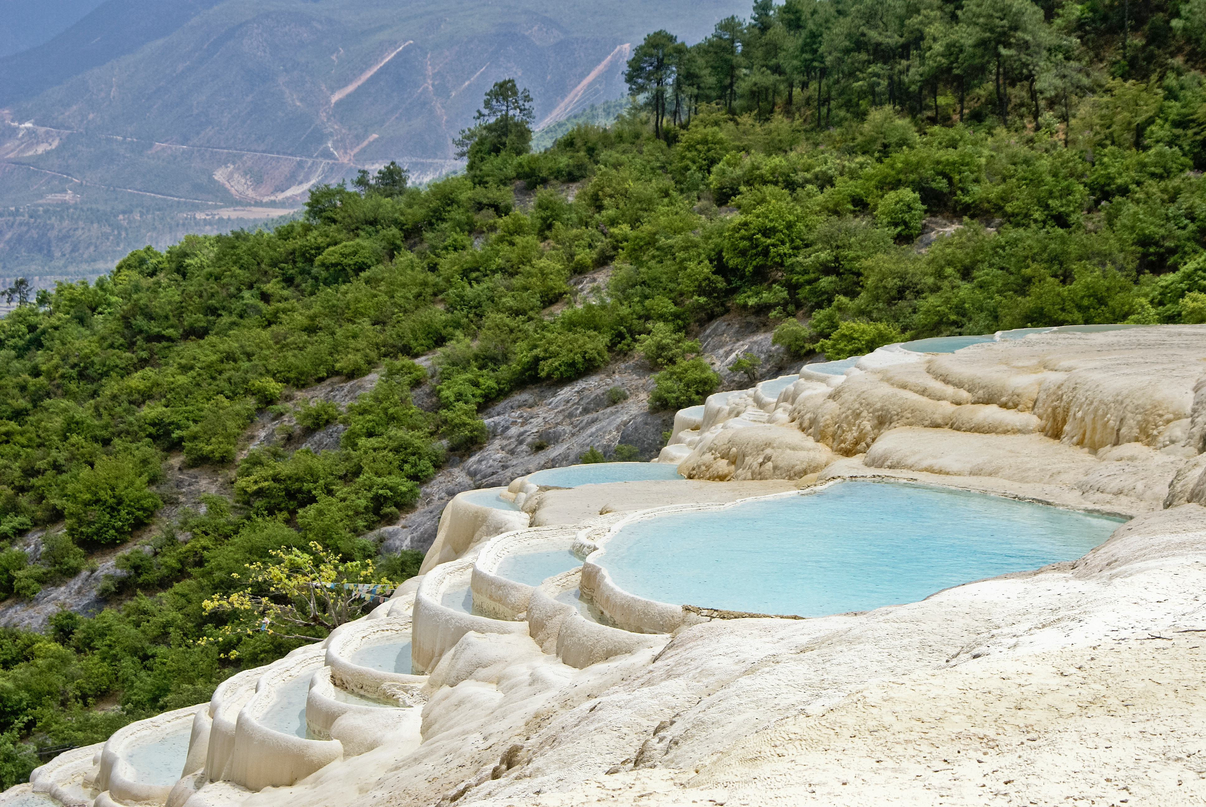 Tiered white travertine pools holding turquoise water at Baishuitai, with forested slopes and distant mountains behind