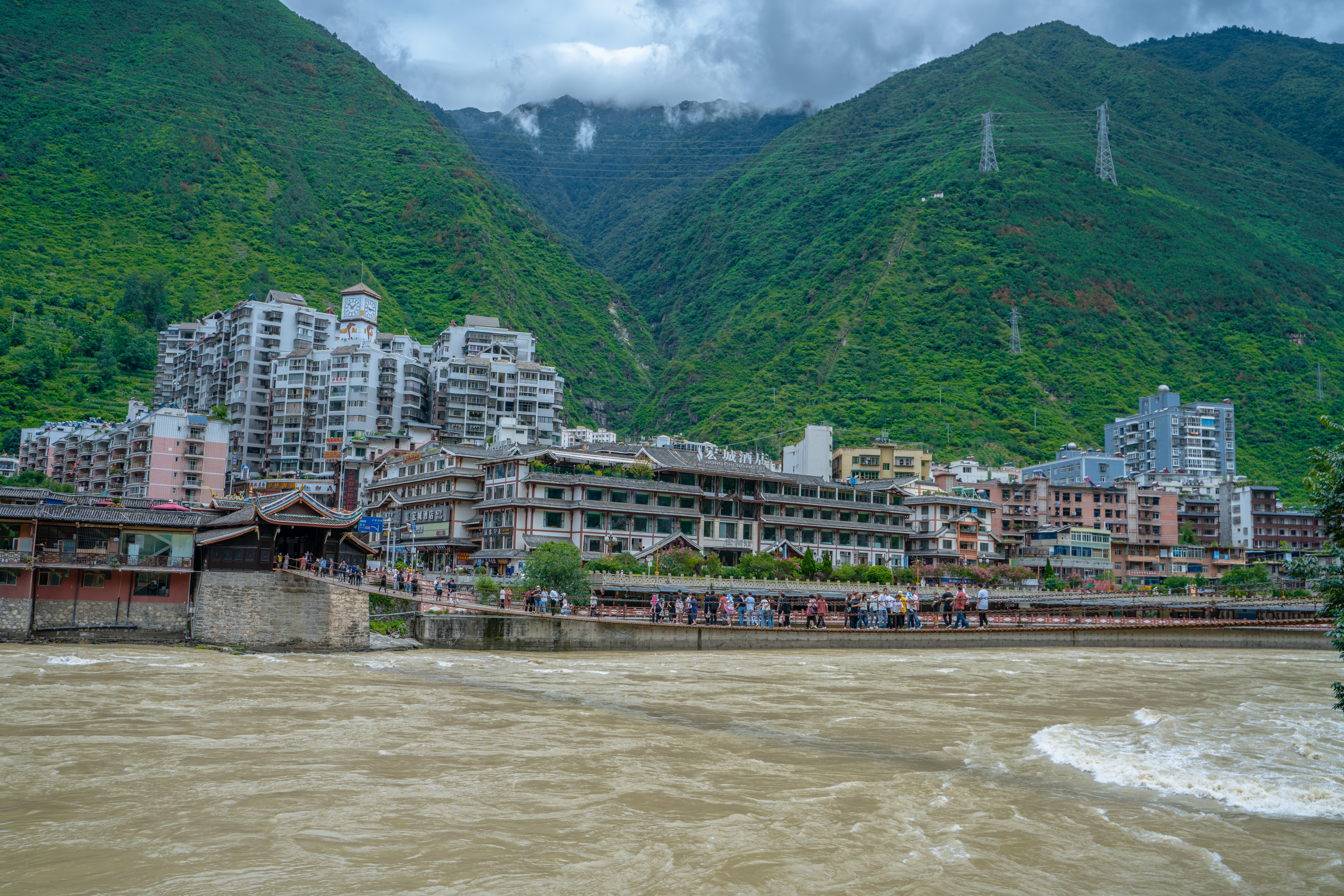 Luding town along the Dadu River in Sichuan, with apartment buildings against green mountains and a pedestrian bridge crossing the muddy water