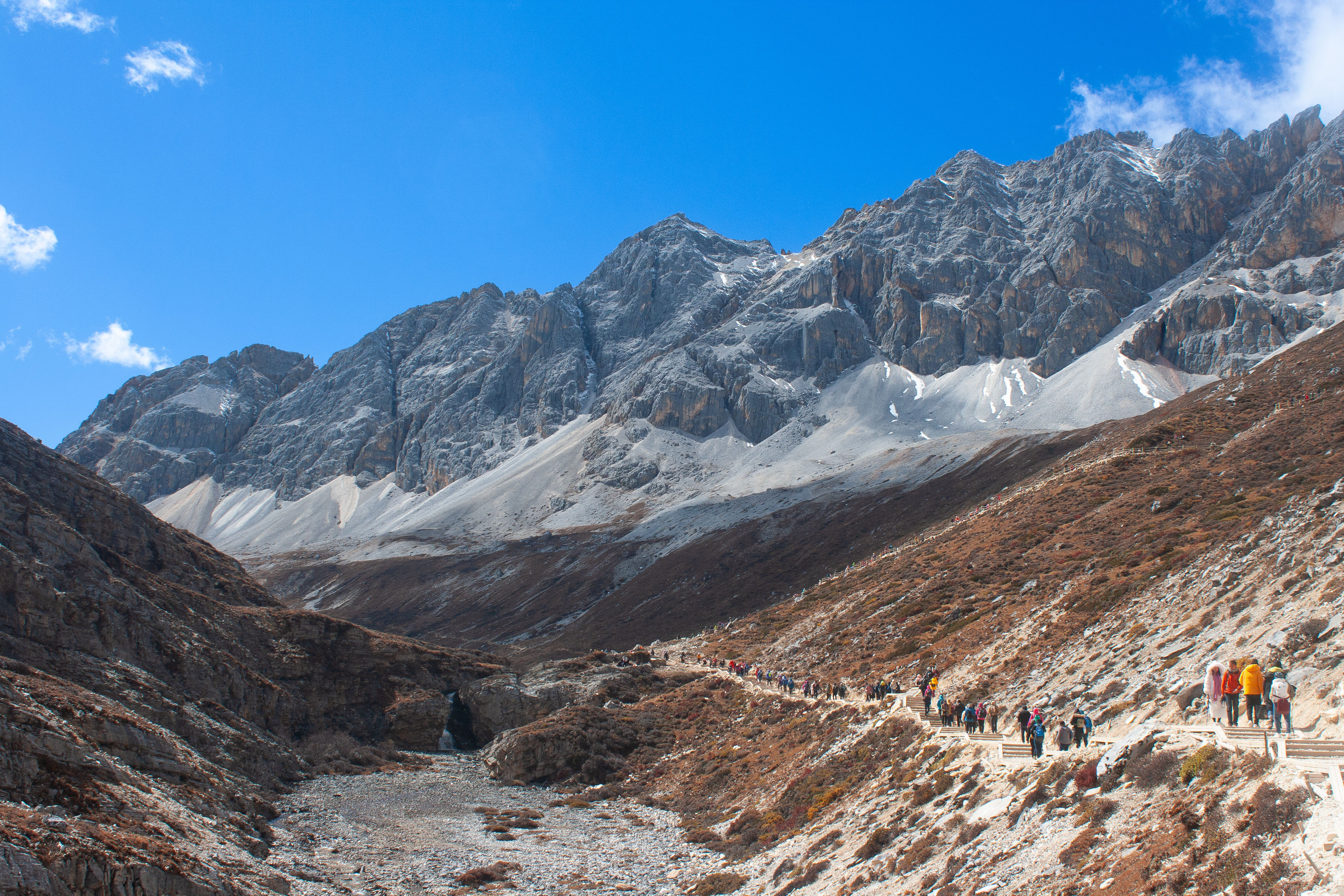 Hikers wind along a stone trail beside a rocky valley floor toward jagged snow streaked peaks in Yading Nature Reserve, Sichuan, China