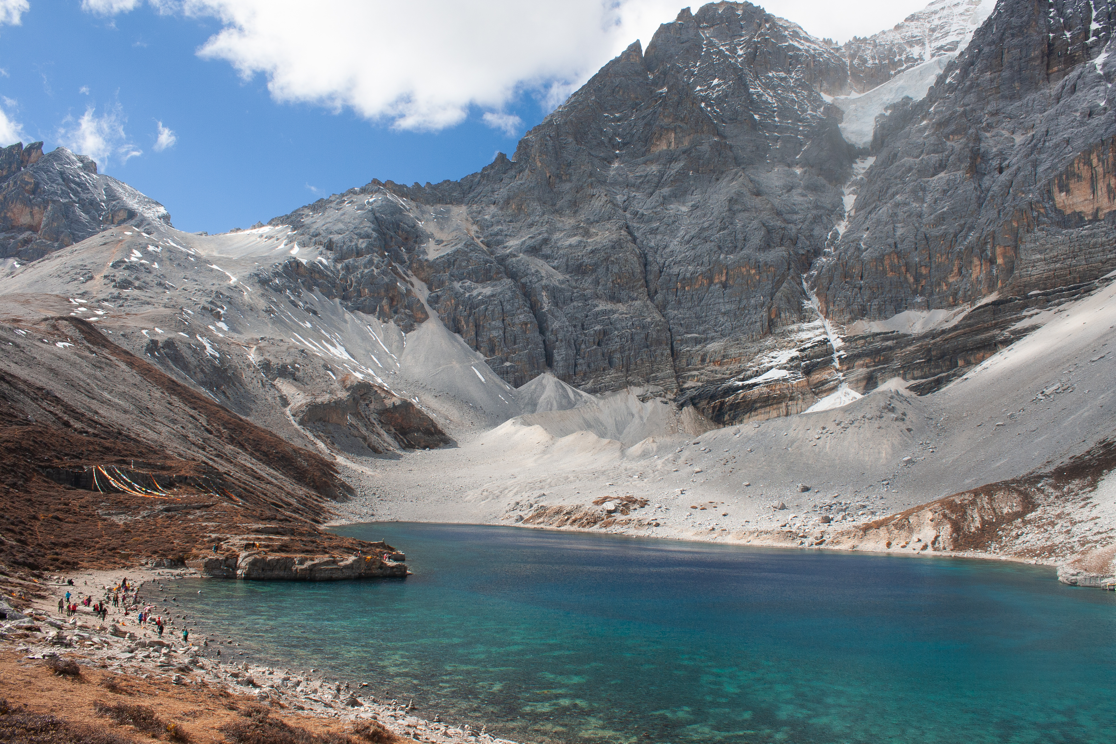 Turquoise glacial lake below jagged snow dusted peaks in Yading Nature Reserve, with trekkers walking along the rocky shoreline