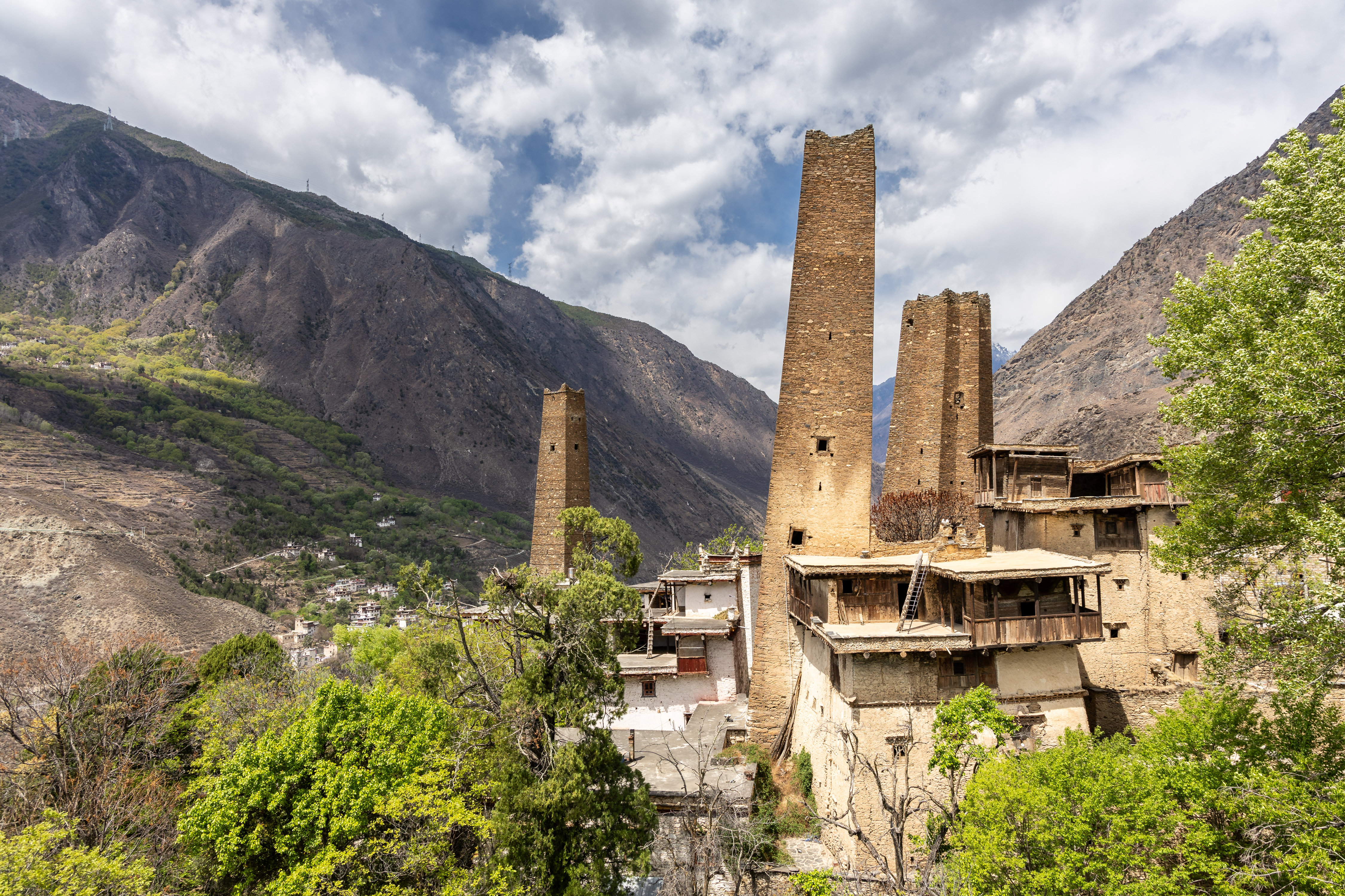 Tall stone watchtowers rise above a fortified stone and timber house in Suopo village, with bare mountain slopes behind, Danba, Sichuan