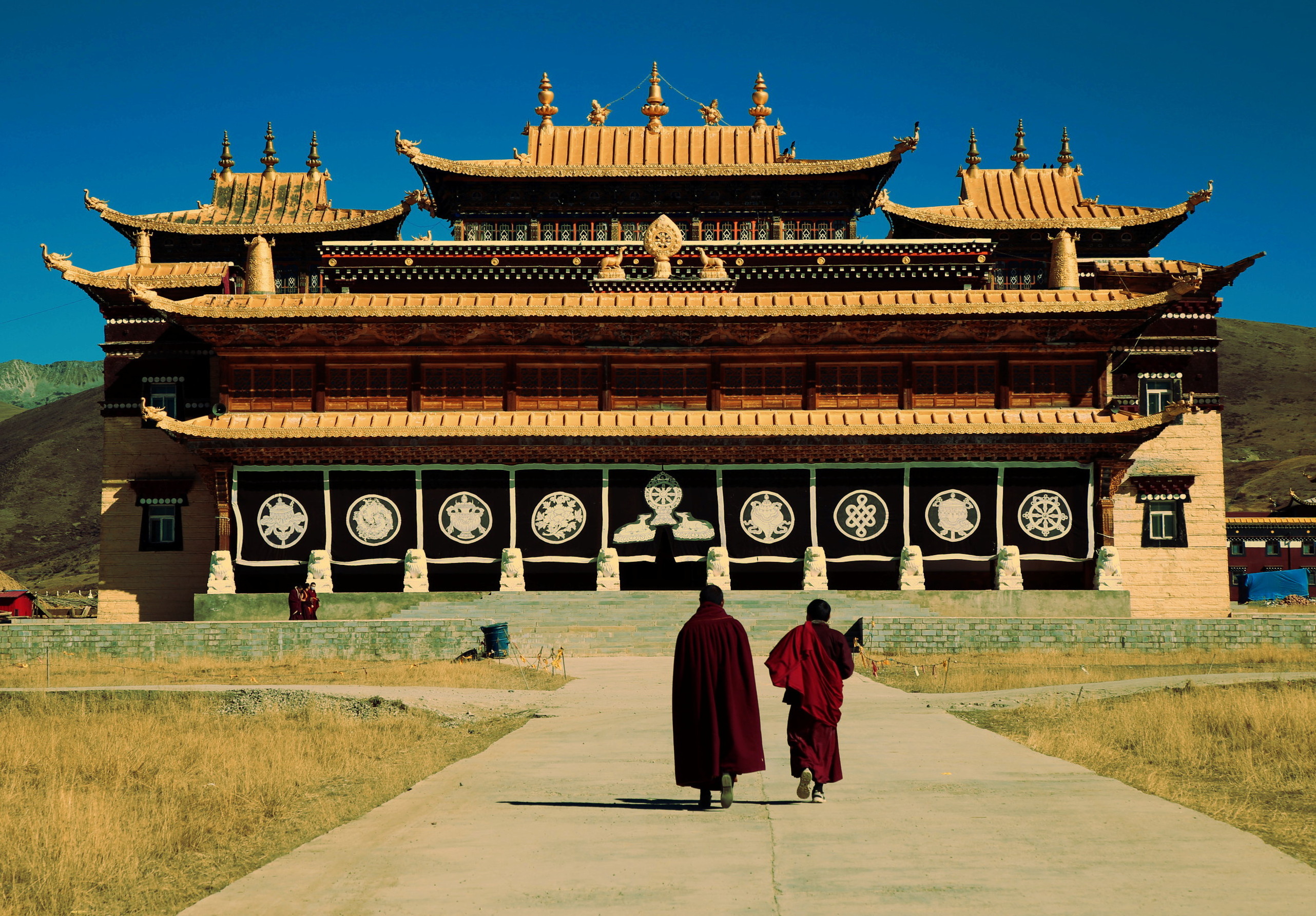 Two monks in dark red robes walk toward the golden-roofed facade of Tagong Monastery under a clear blue sky in western Sichuan