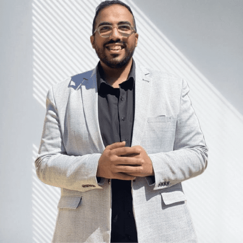 Man in a light gray blazer and black shirt standing with hands clasped, smiling at the camera.