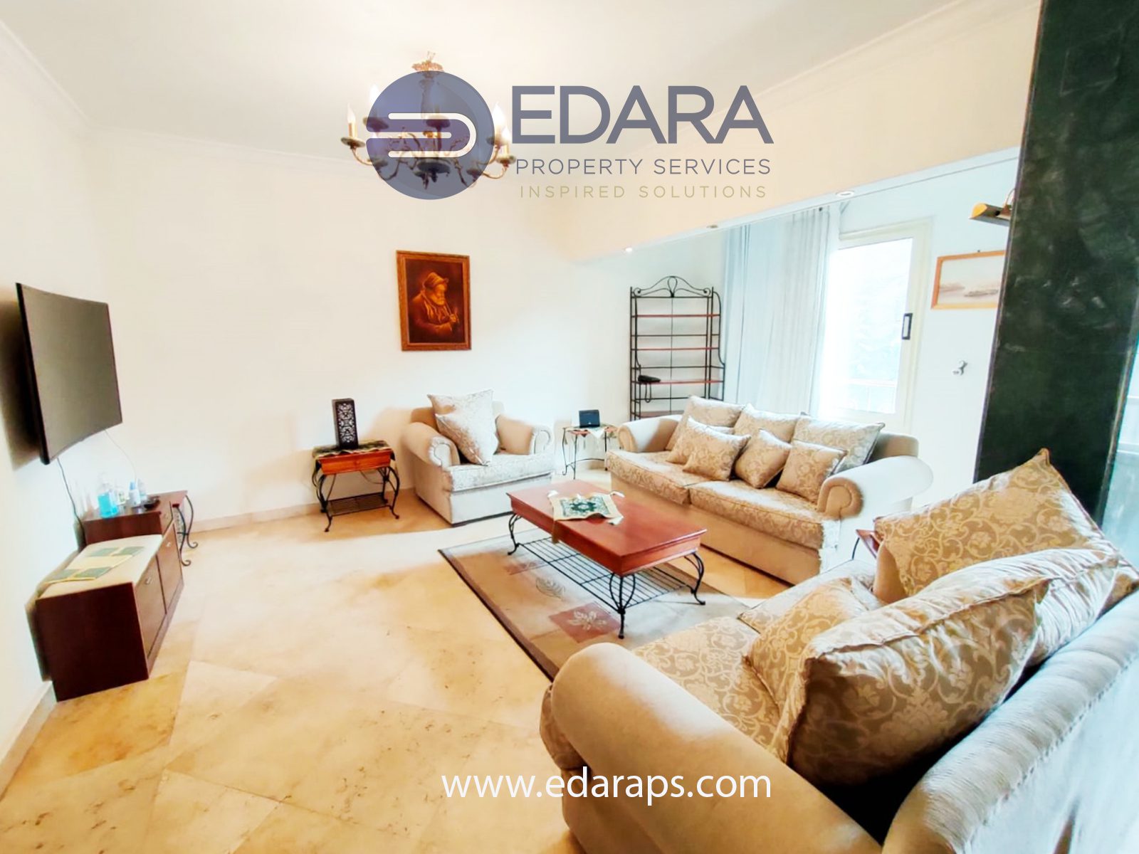 Living room with beige sofas, a coffee table, and a television mounted on the wall in a well-lit space.