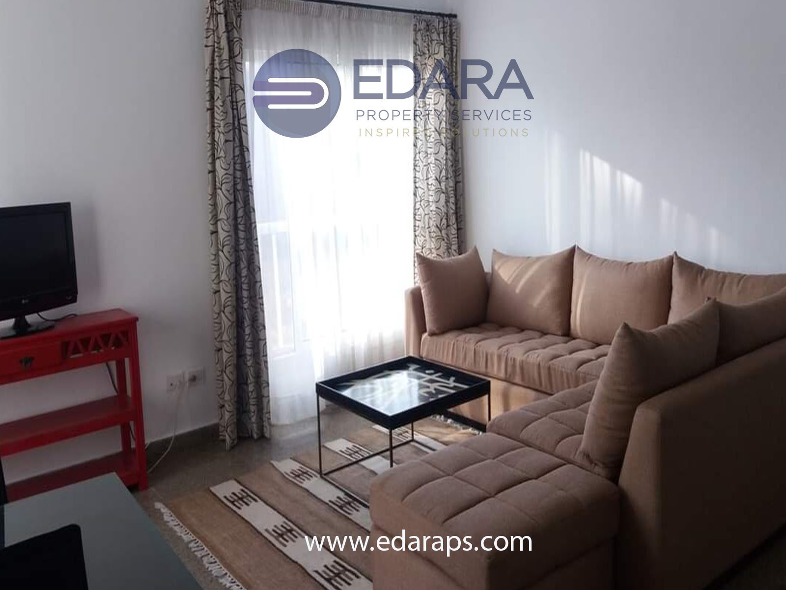 A modern living room featuring a beige sofa, glass coffee table, and television set against a light backdrop.