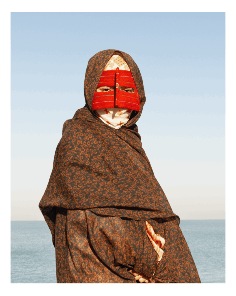 A woman fully wrapped in a brown garment stares directly at the camera. She is wearing a red mask that covers her face such that only her eyes are visible.