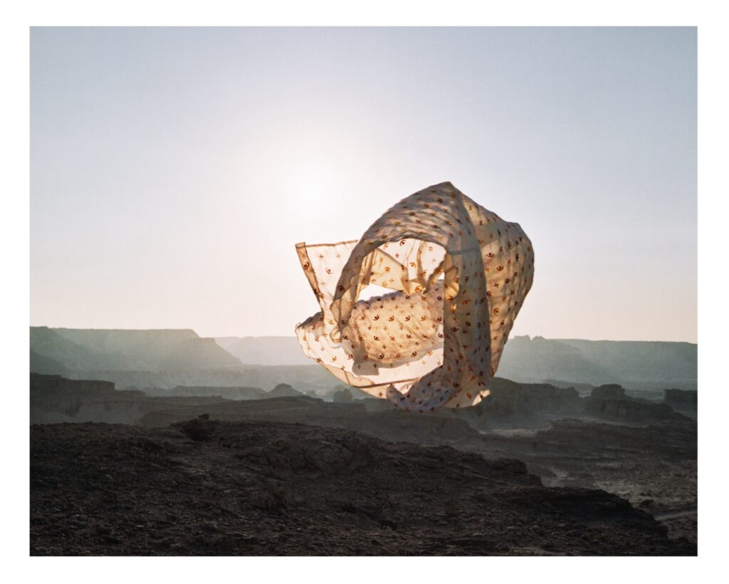 A large piece of fabric is skyborne - spread out in cylindrical shape. Beneath, a rocky landscape.